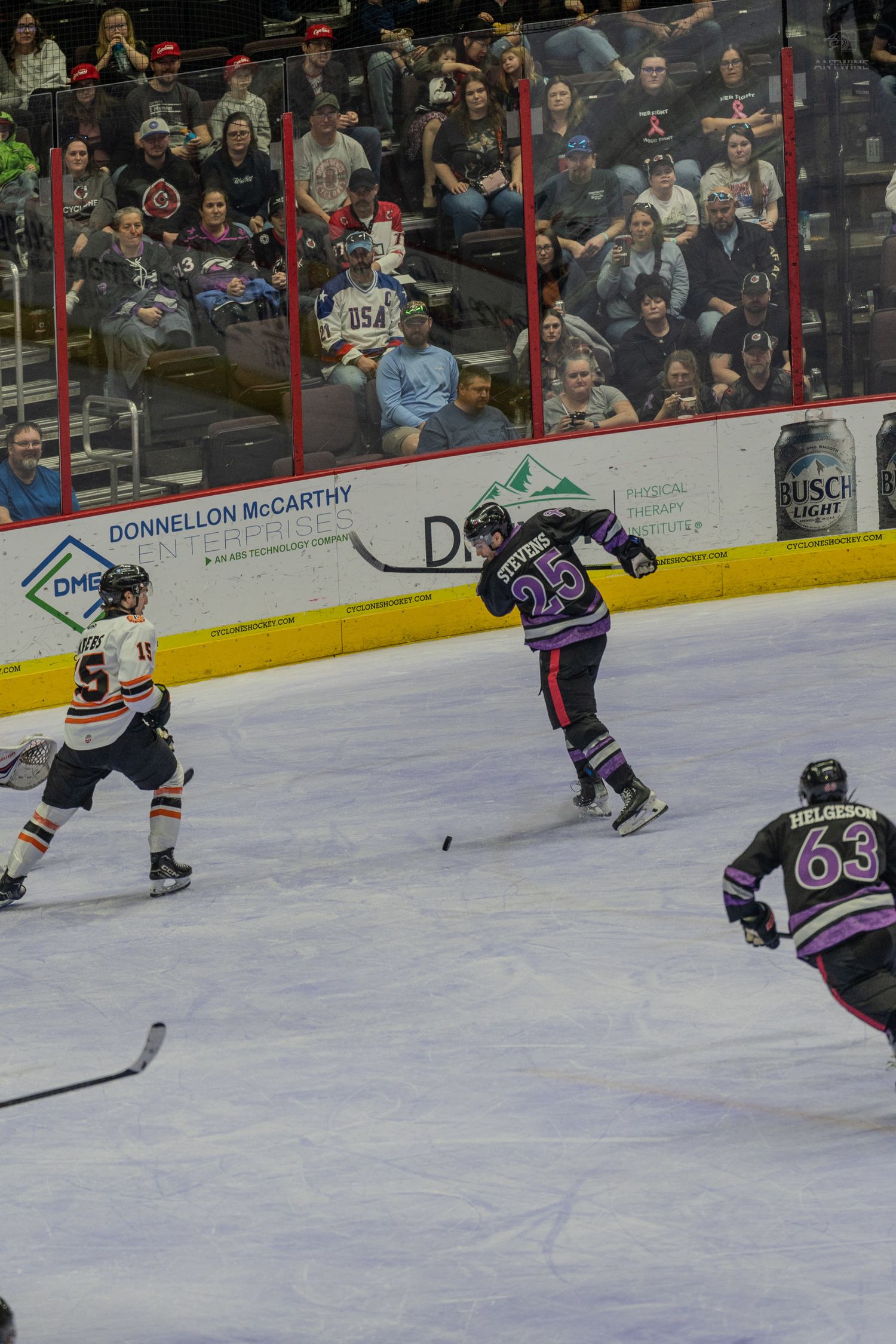 Hockey player #25 in purple jersey shoots puck on ice rink during professional hockey game with fans watching.