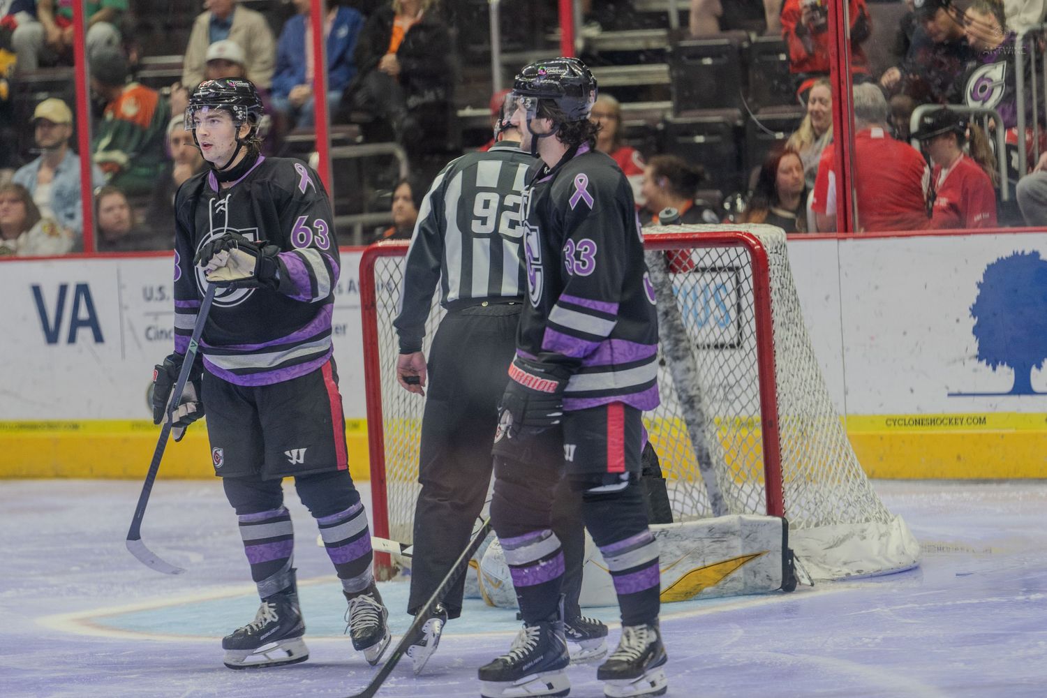Two hockey players in purple jerseys stand near the goal with referee number 92.