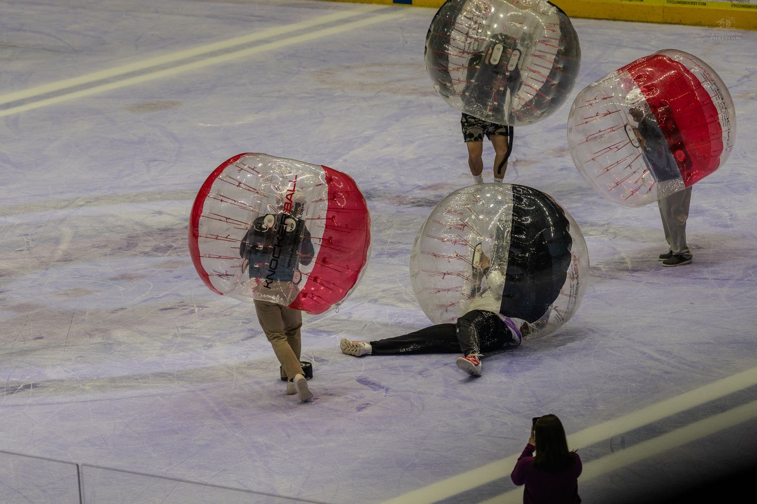 Four players in red and black bubble soccer balls fall on an ice rink during an intermission game.