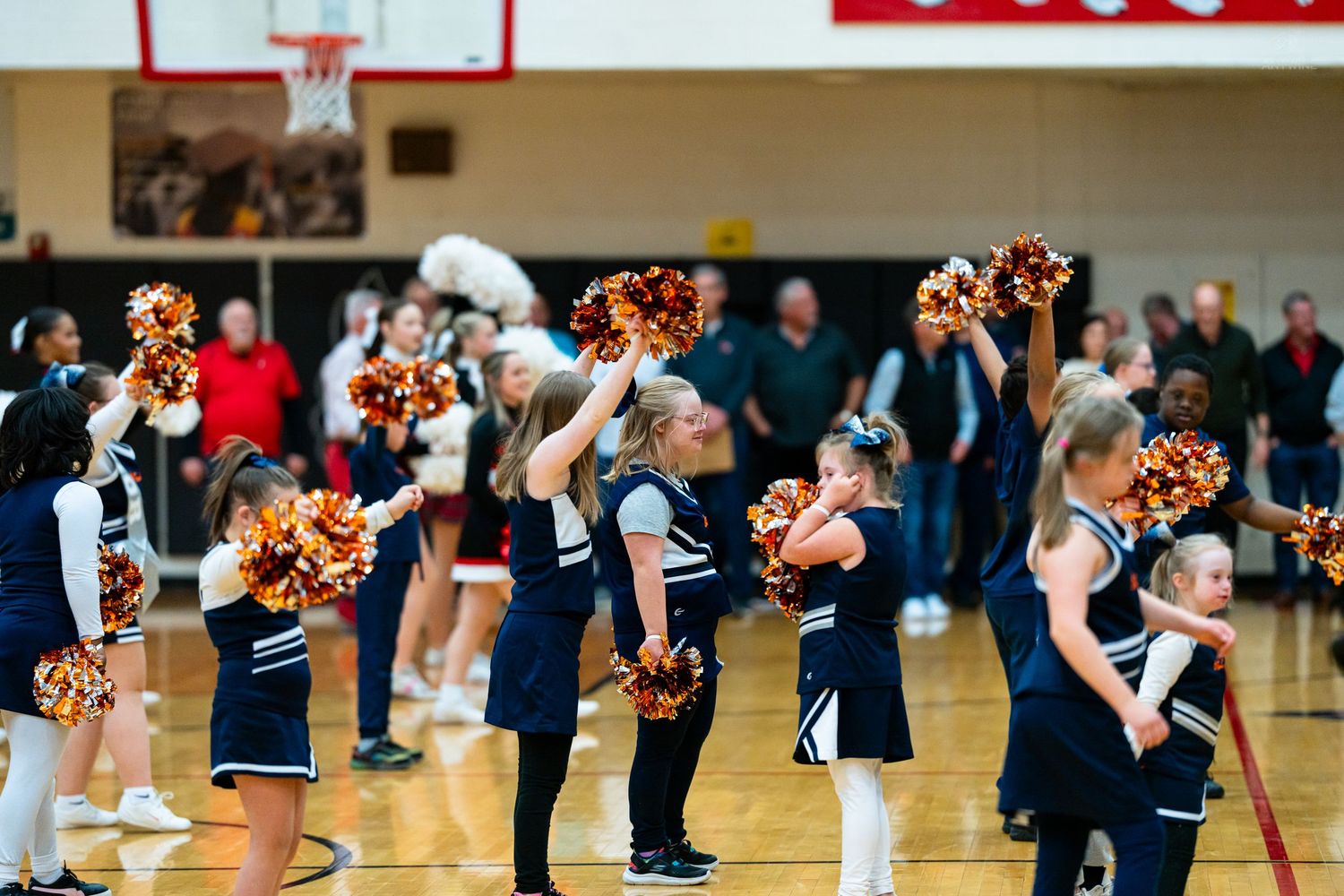 Young cheerleaders in navy uniforms hold gold and burgundy pom-poms up during a basketball gym performance.