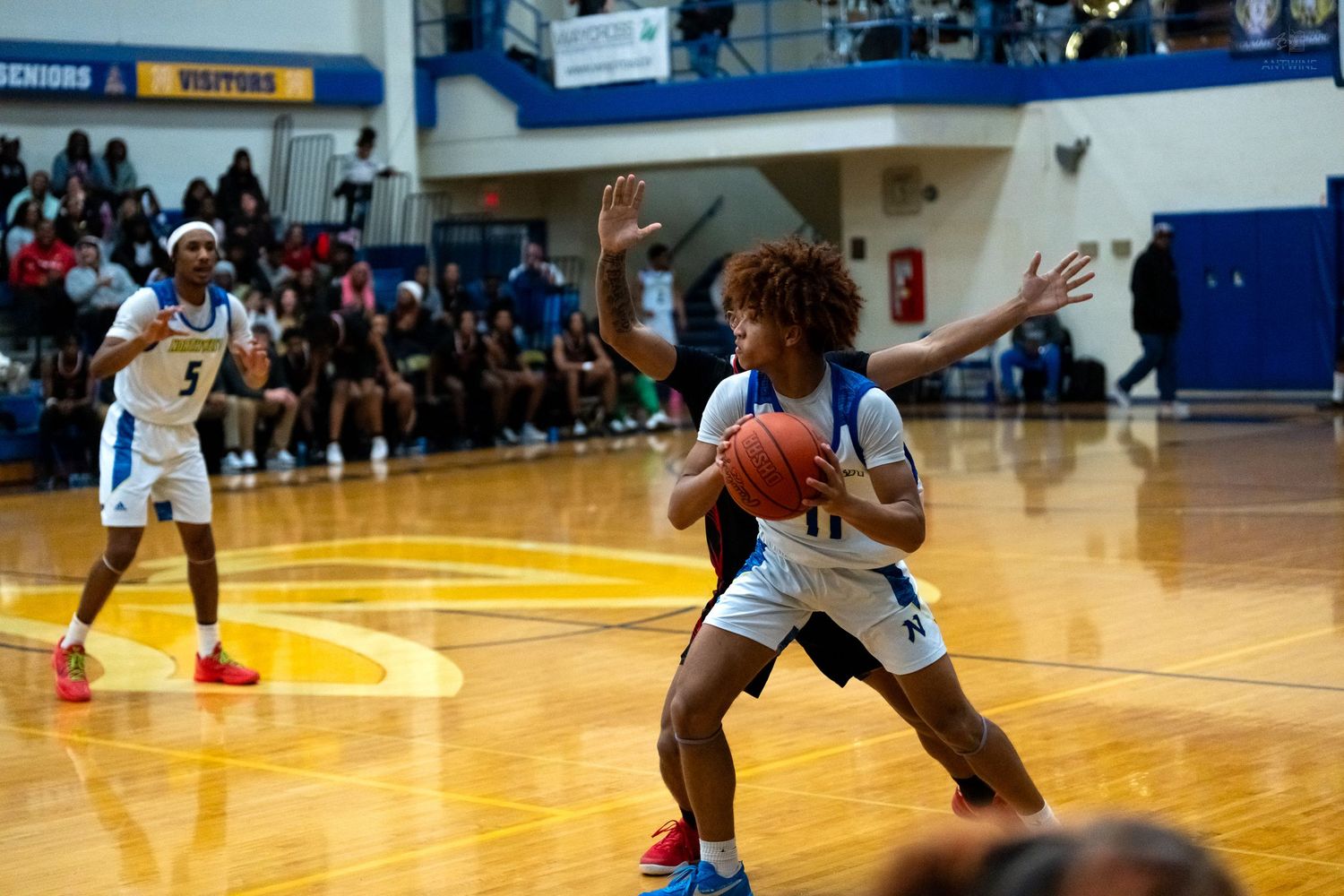High school basketball player in blue uniform pivots with the ball while defender reaches in during a gymnasium game.
