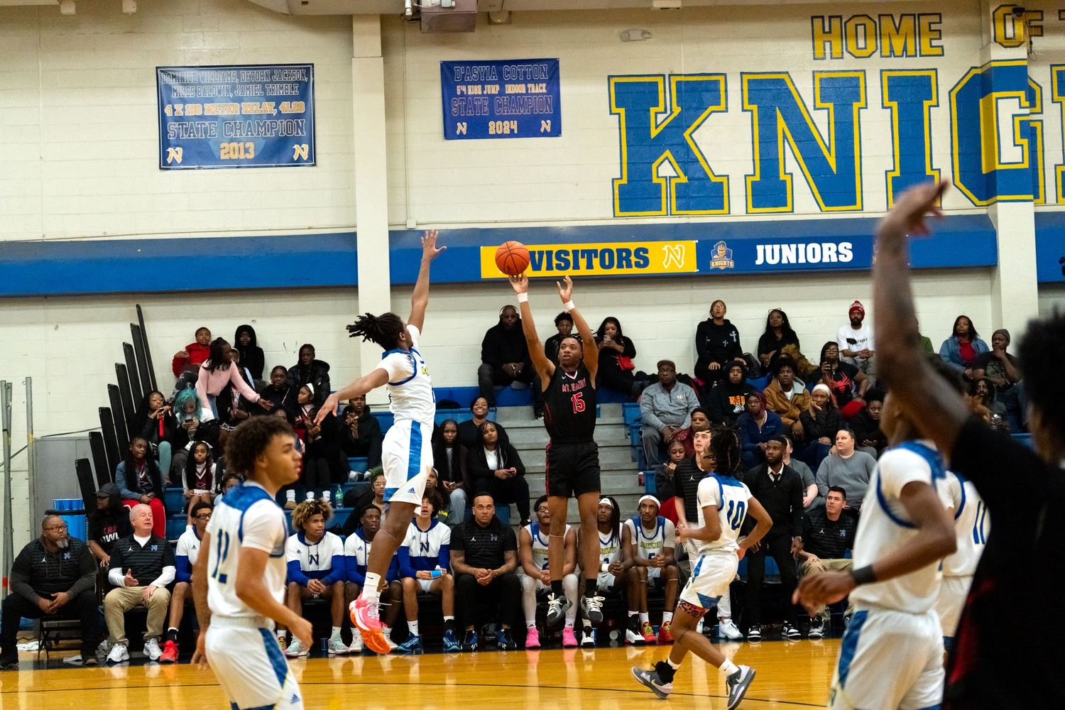 Basketball player shoots a jump shot in a high school gym with HOME KNIGHTS signage visible.