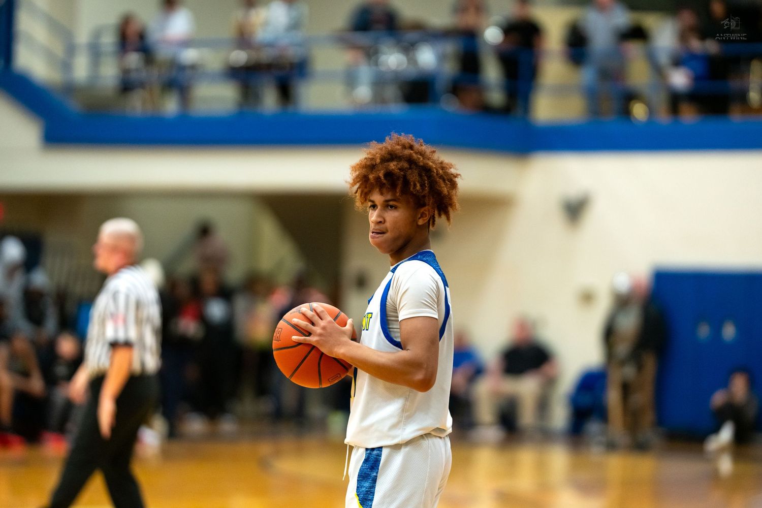 Curly-haired high school basketball player in white uniform holds a basketball on a blue gymnasium court.