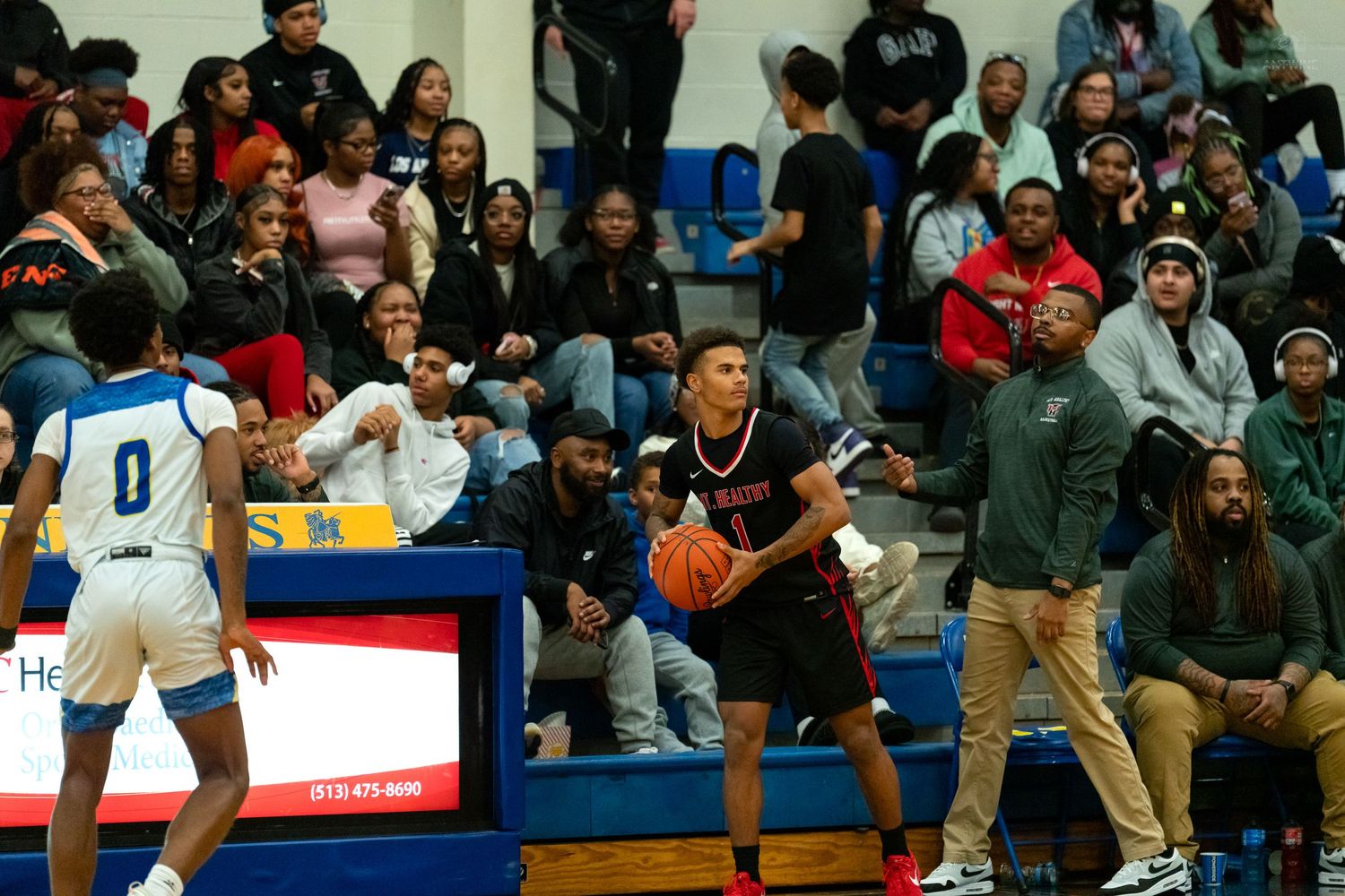 High school basketball player holds the ball near the sideline during a game in a crowded gymnasium.