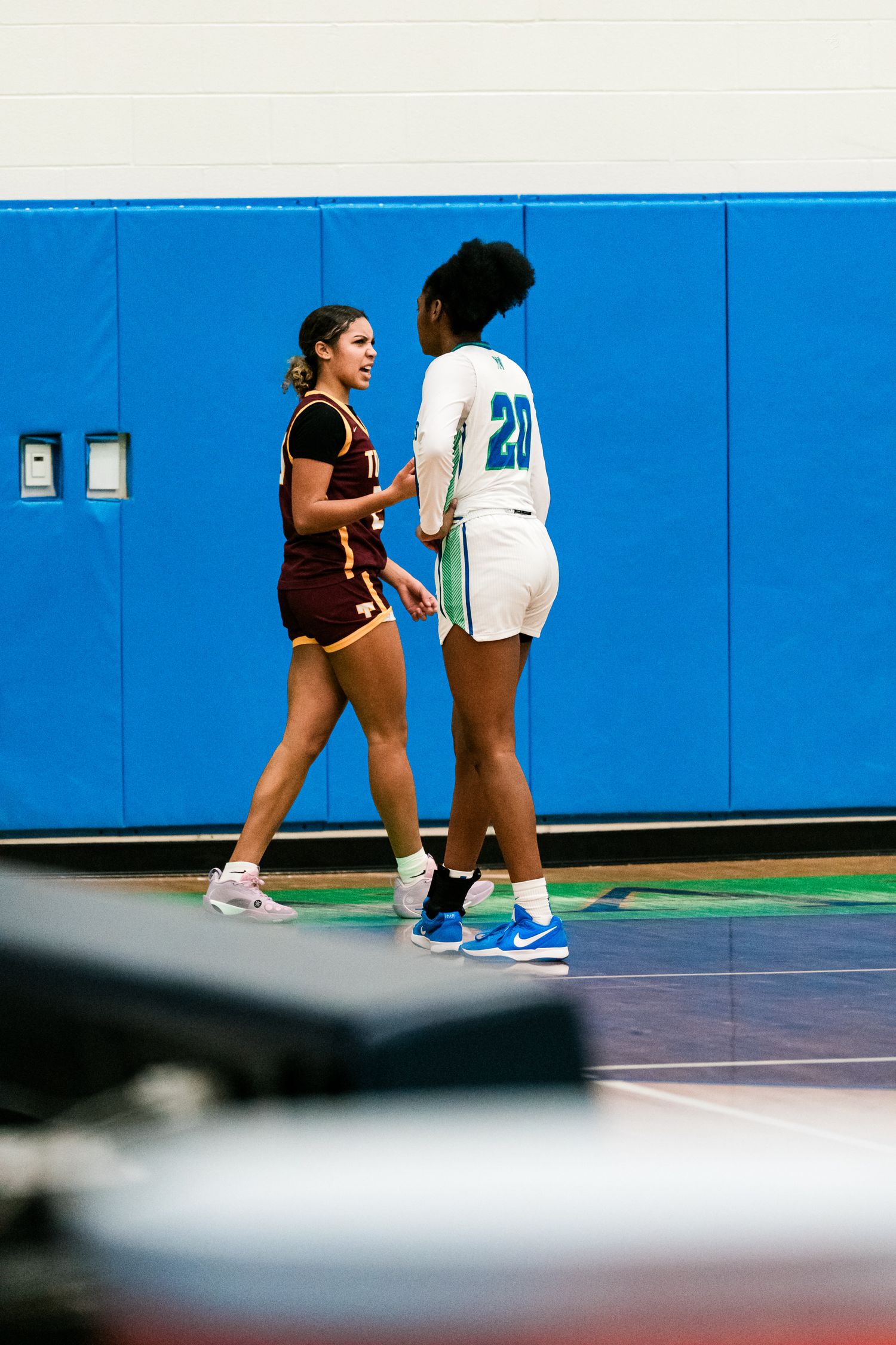 Two female high school basketball players in maroon and white uniforms face each other near the sideline during a game.