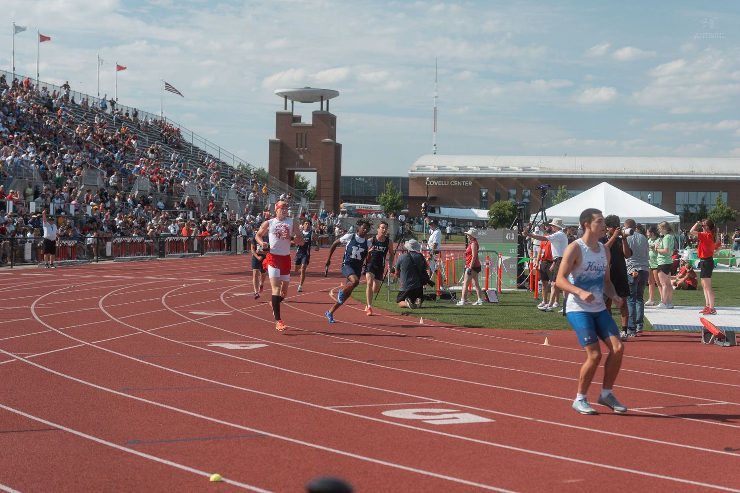 Runners compete on a red track at a large outdoor high school track and field meet with packed stands.