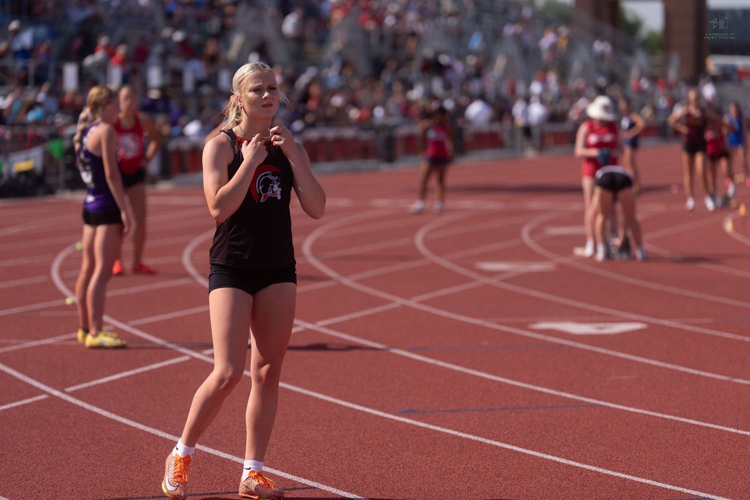 Blonde female track athlete in black uniform claps hands on a red track during an outdoor meet.