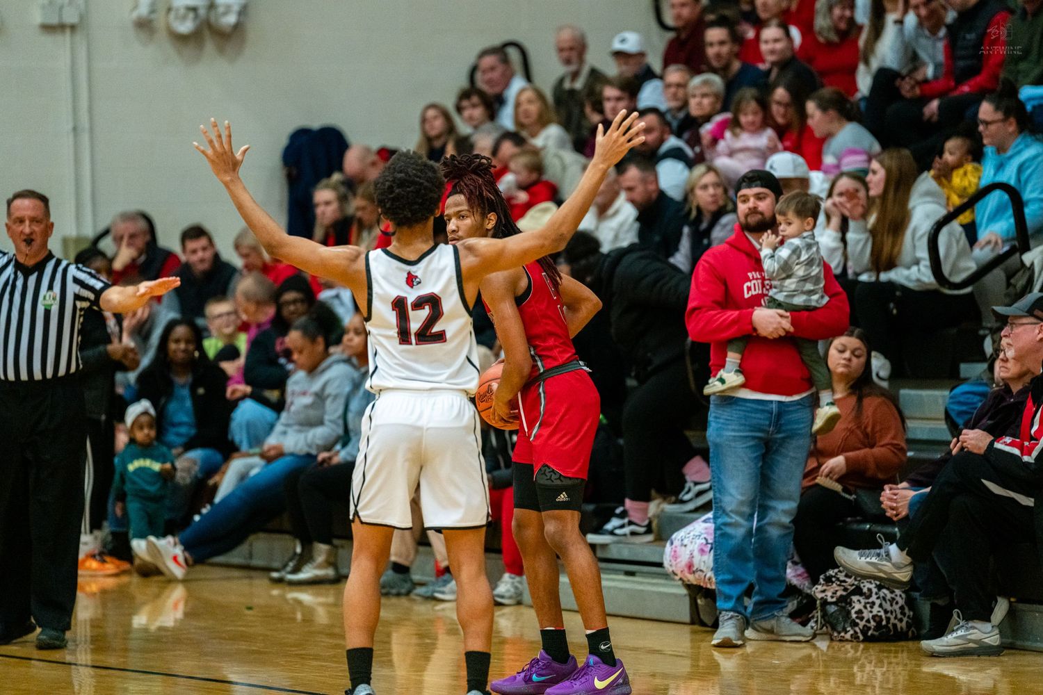High school basketball player #12 raises hands as crowd watches during packed gym game.