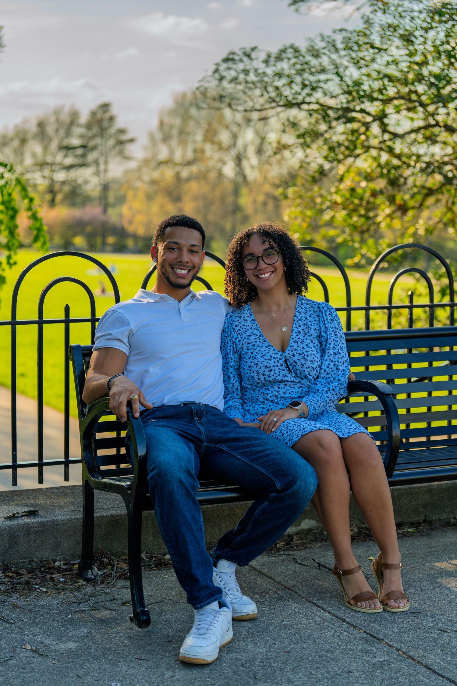 Smiling couple sitting together on a black park bench surrounded by lush green trees.