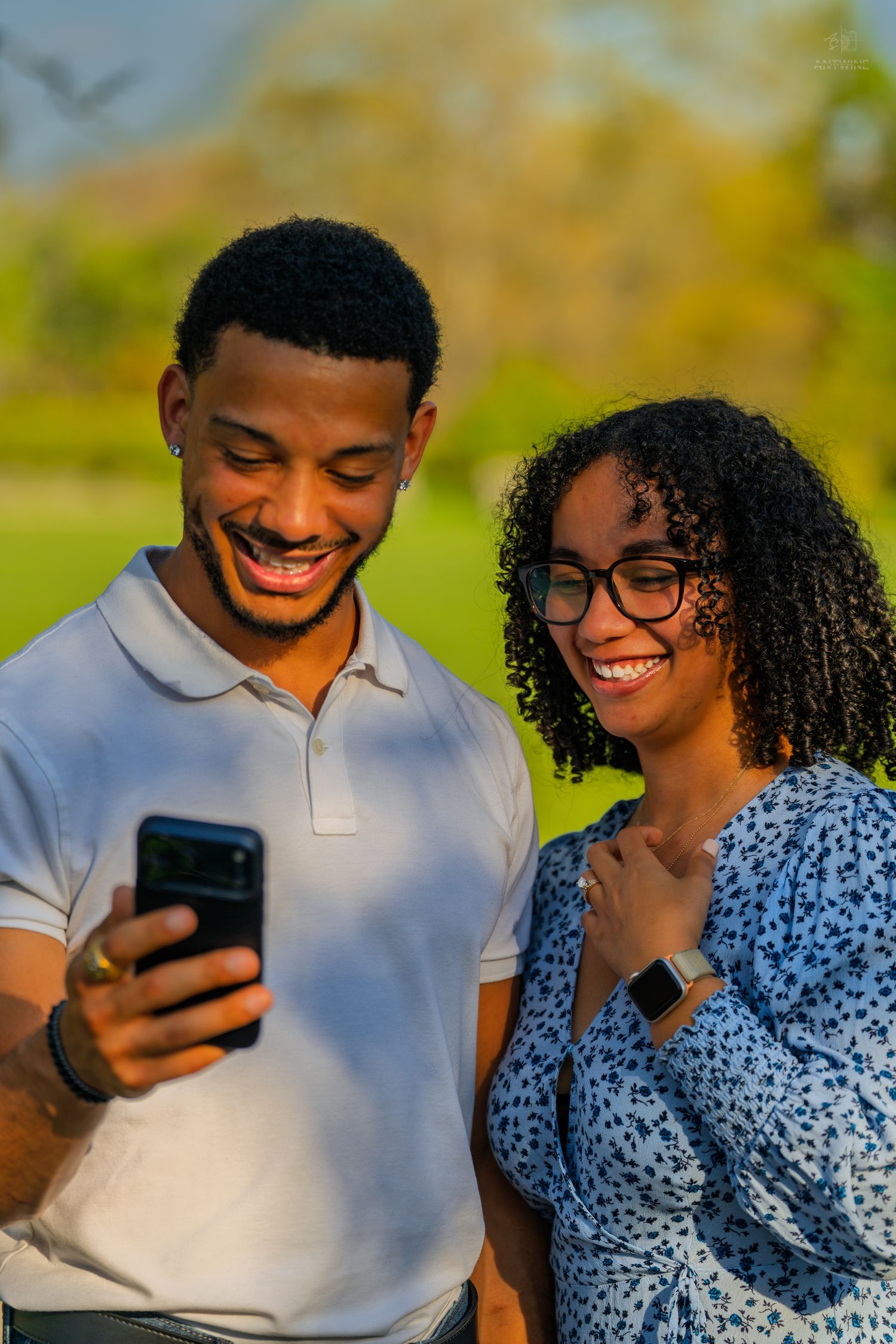 Smiling couple in sunlit park reviewing photos on smartphone, woman wearing glasses and blue floral dress.