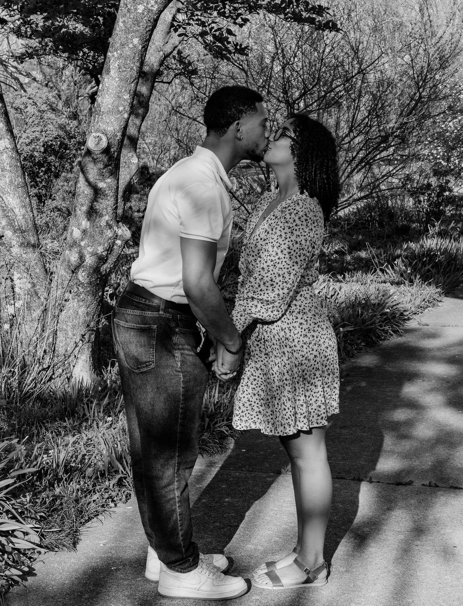 A couple sharing a romantic kiss while holding hands on a sunlit park path surrounded by trees.
