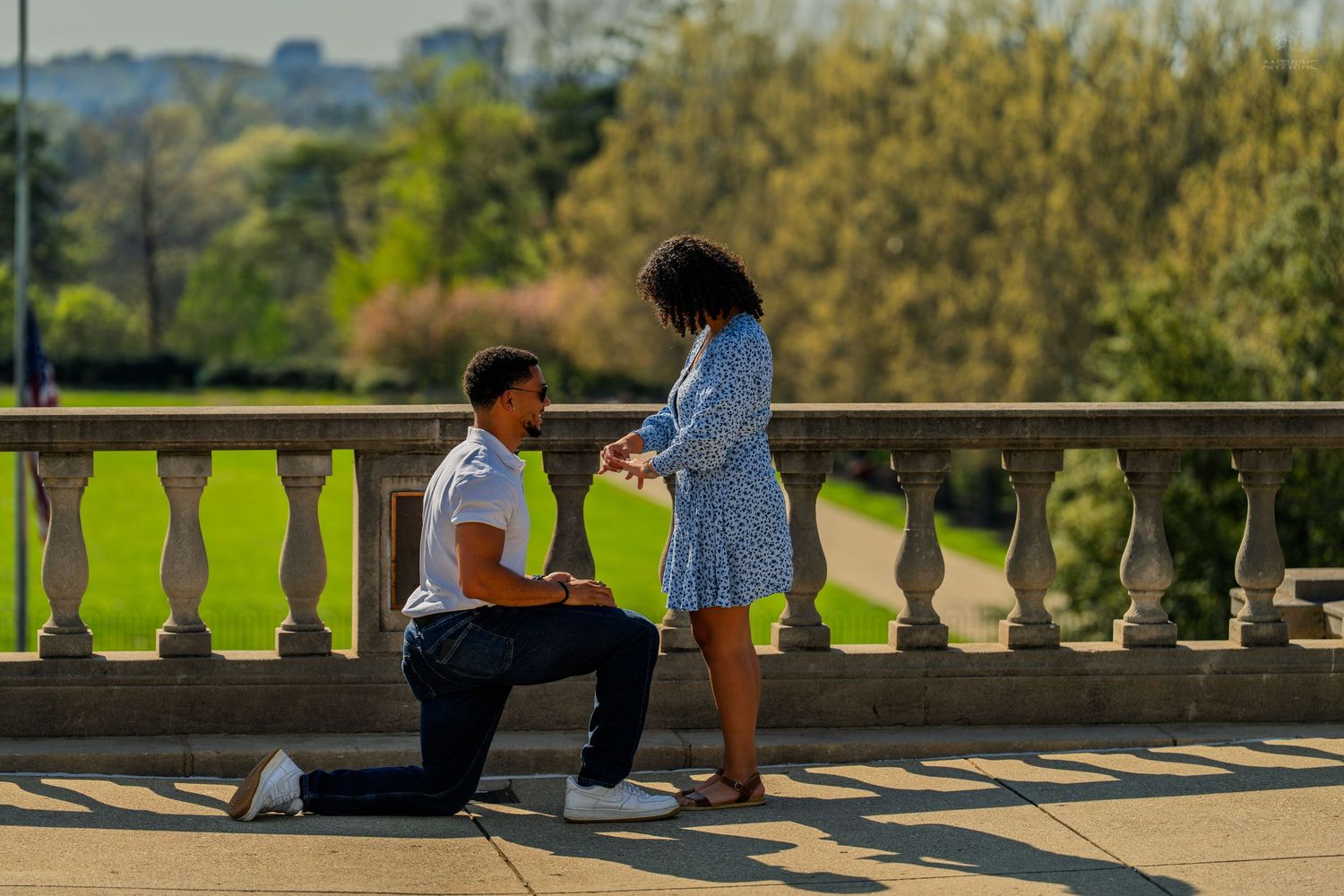 Couple sharing a romantic marriage proposal moment on a sunlit terrace overlooking a green park.