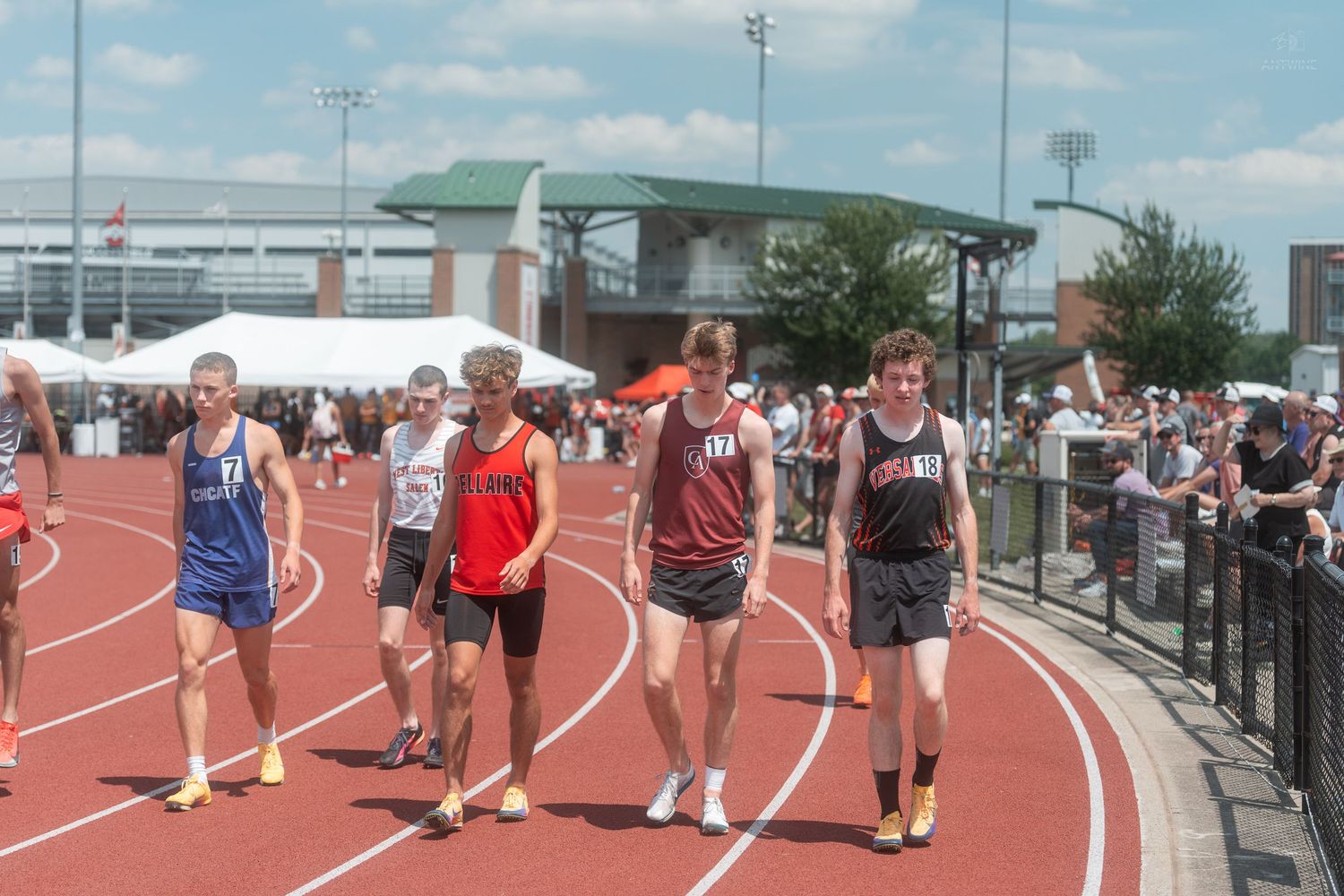 Male track and field athletes walk on a red track at an outdoor stadium during a sunny competition.