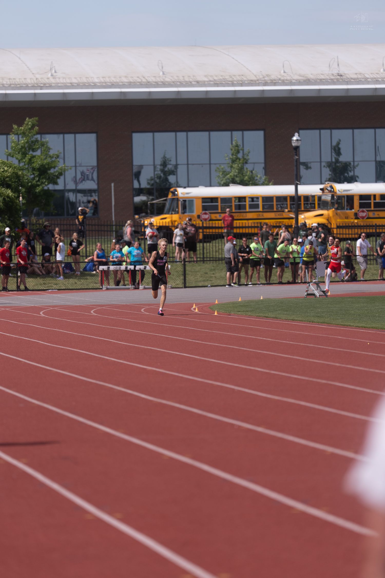 A lone athlete runs on a red track at a high school track meet with yellow school buses in background.