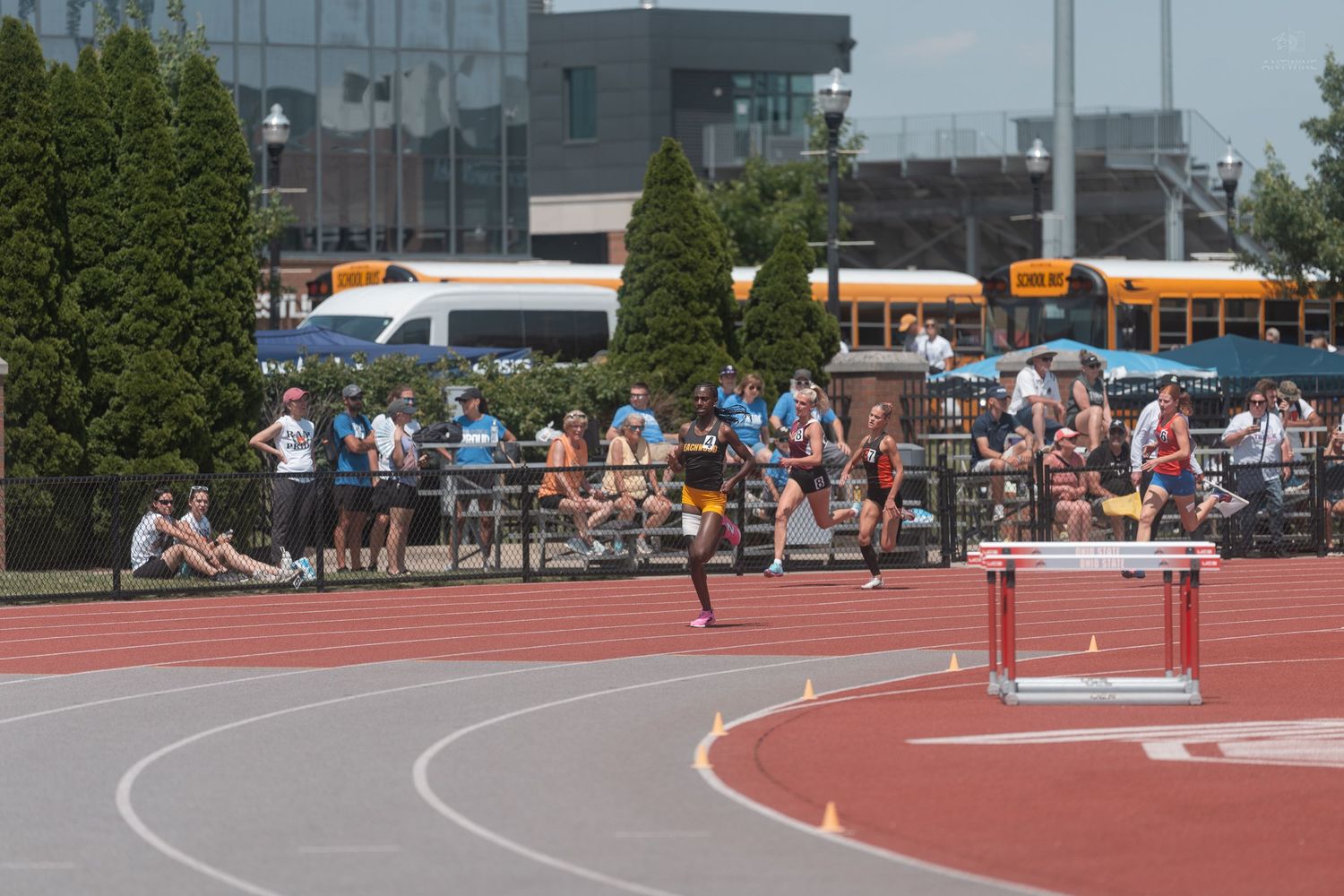 Track athletes racing past a hurdle on red track during outdoor championship meet.