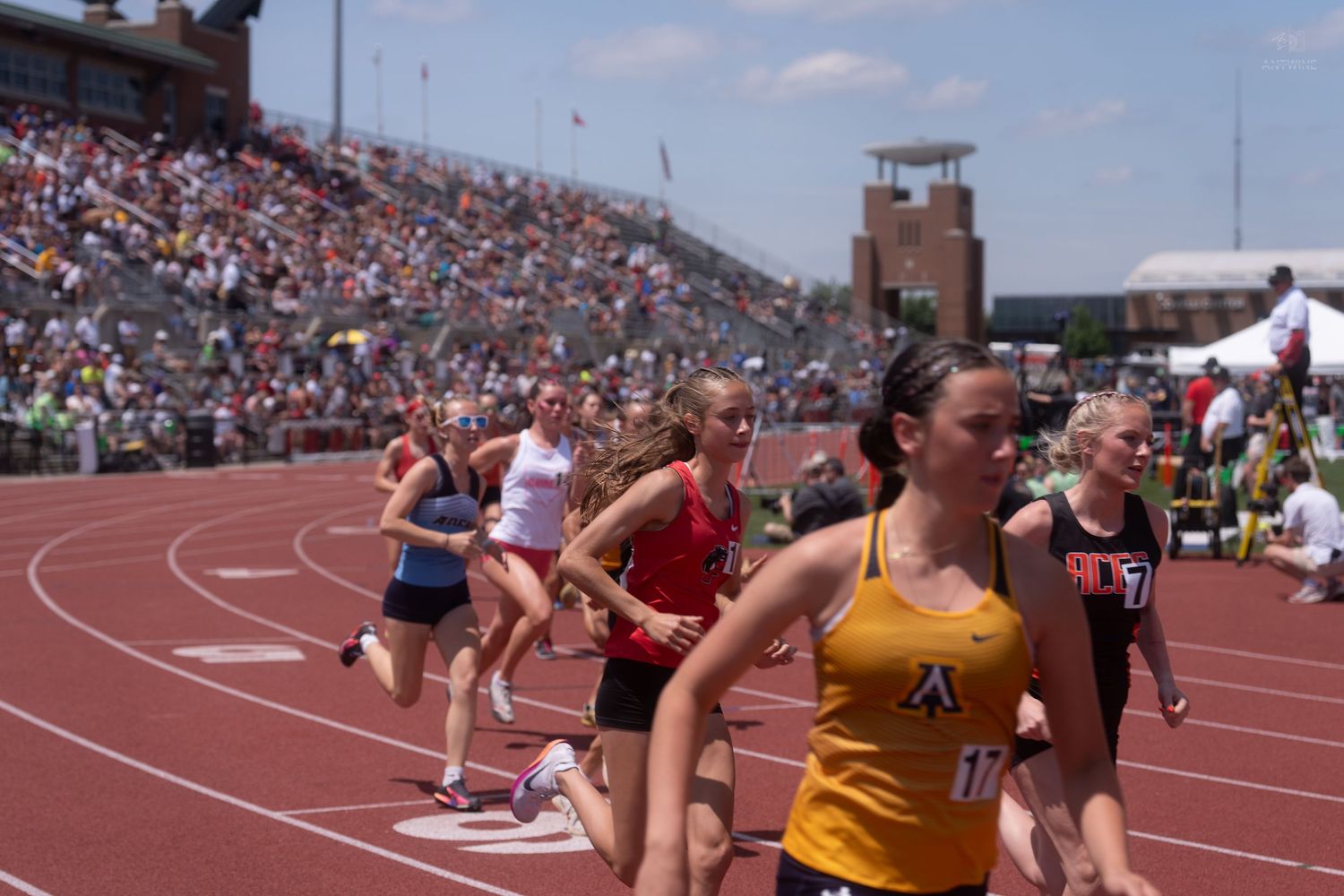 Female track athletes compete in a relay race at a packed outdoor stadium on a sunny day.