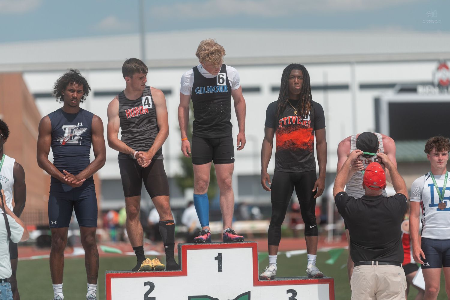 High school boys stand on numbered award podium at sunny OHSAA track and field championship meet.
