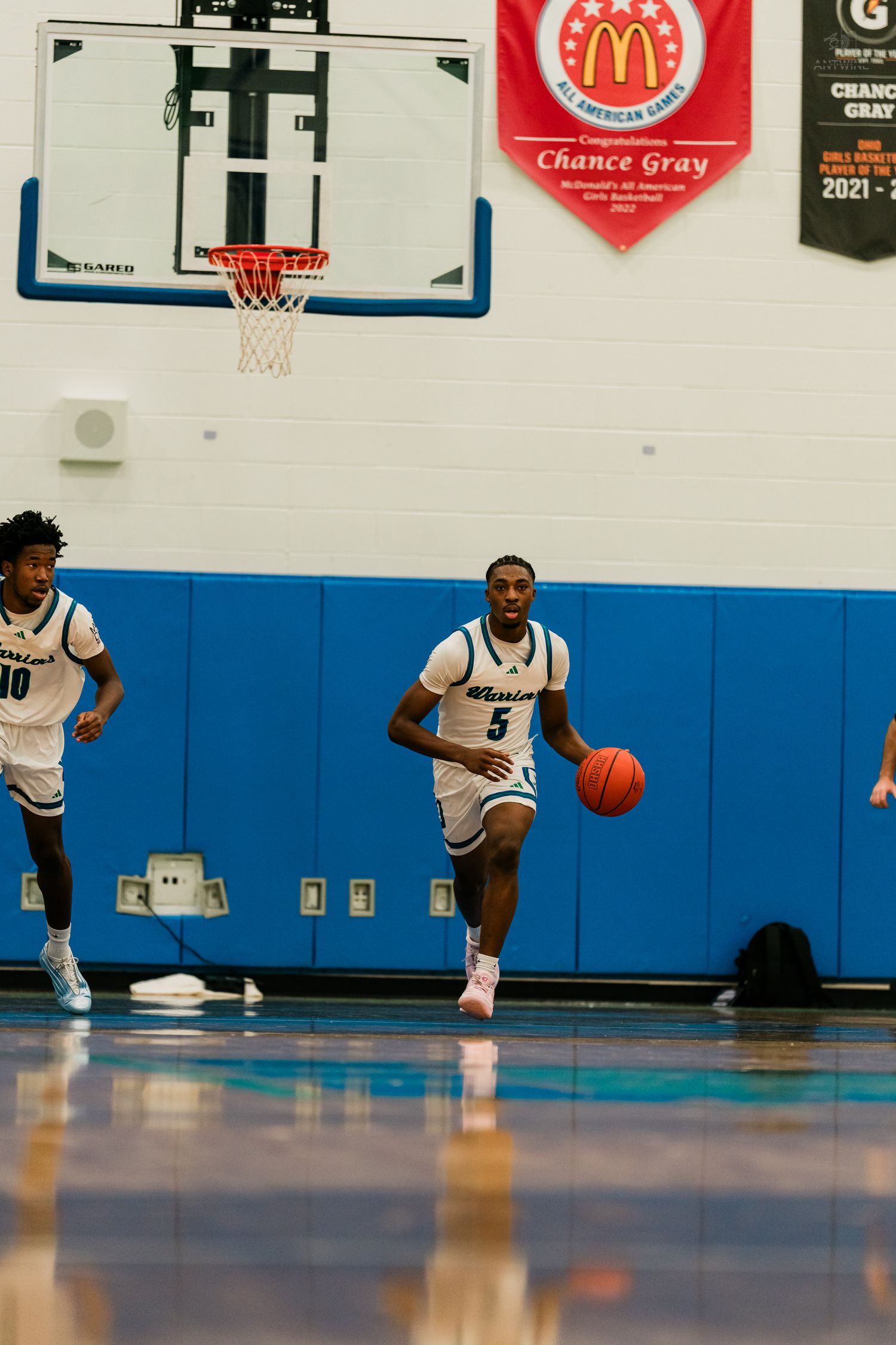 High school basketball player #5 in white Warriors uniform dribbles down the court during a game.