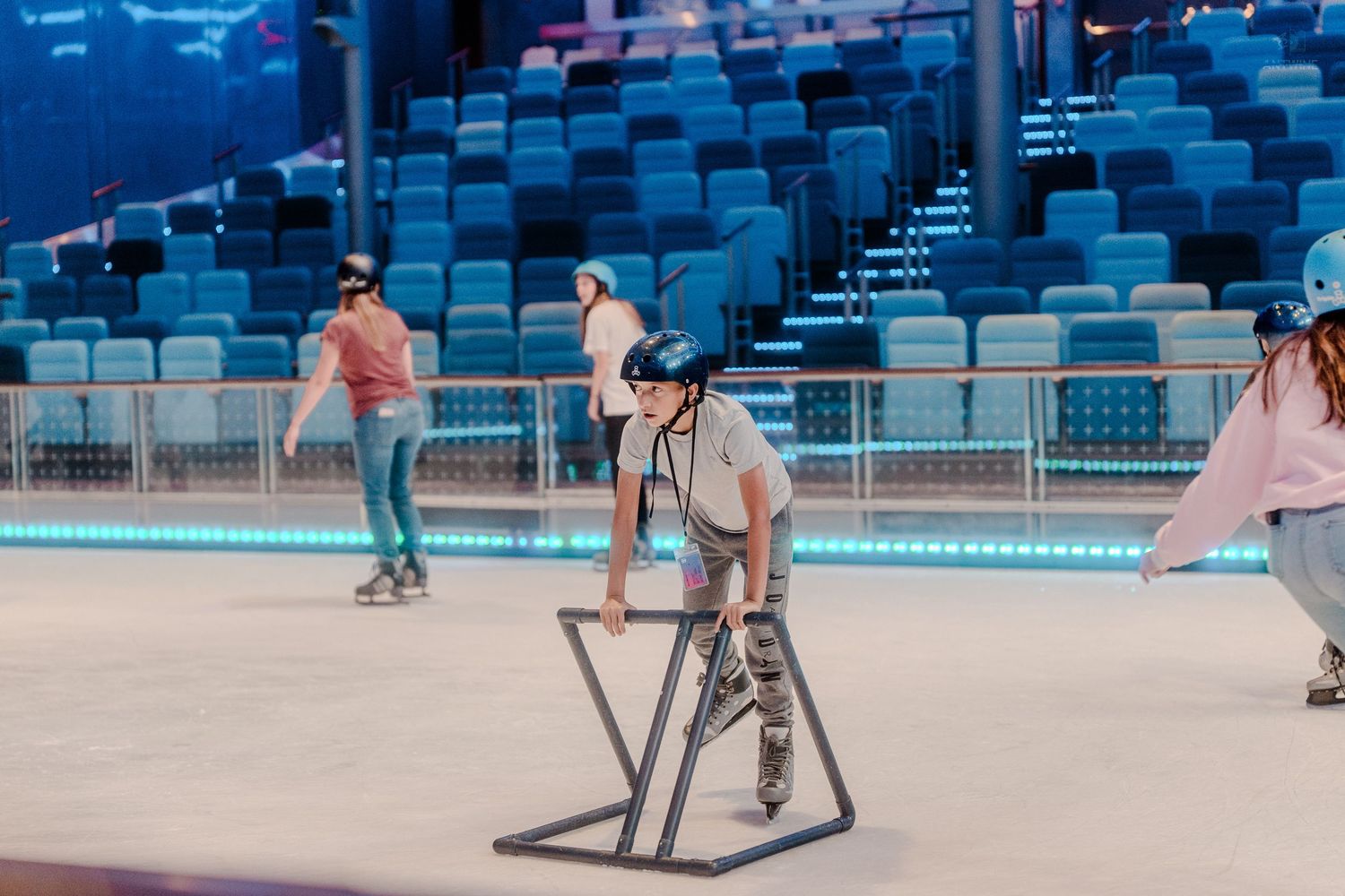 A child in a helmet uses a metal skating aid while learning to roller skate on a cruise ship indoor rink.