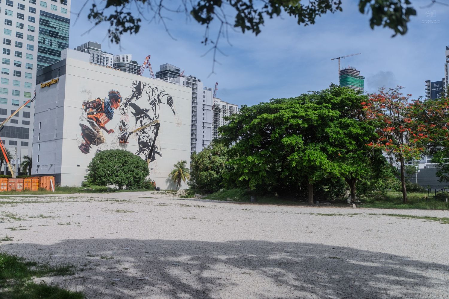 Large street art mural of figures on a white building wall beside an open lot with a flowering tree.