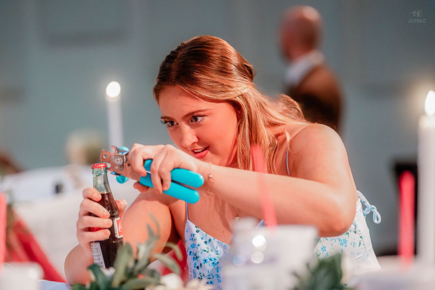 Blonde woman using a bottle opener at an elegantly decorated wedding reception table with candles.