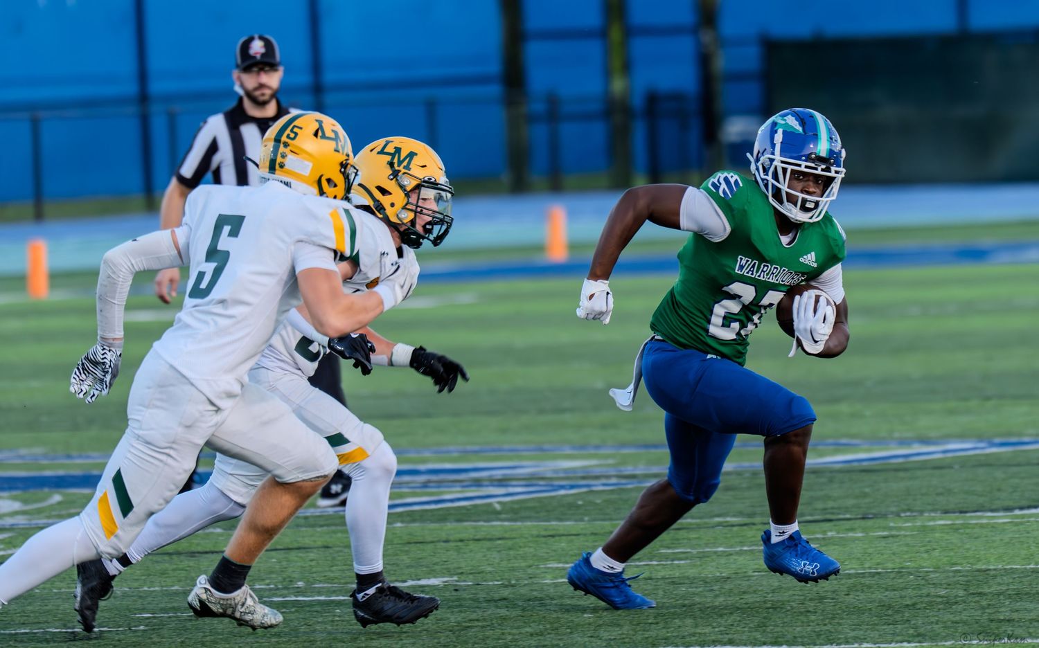 Warriors #26 running back in blue and green uniform rushes past defenders during game.