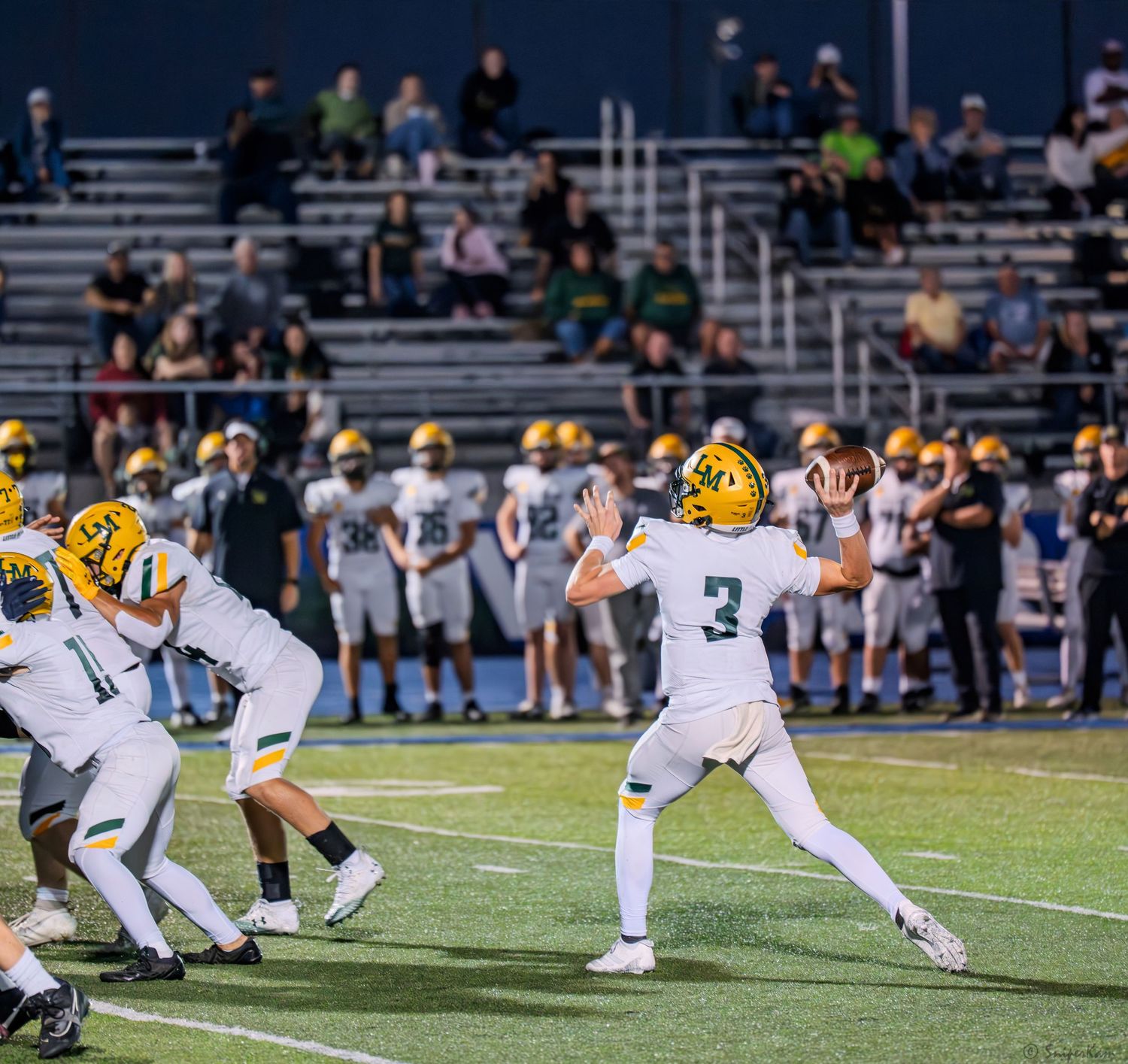 A quarterback in gold helmet throws a pass during a high school football game under stadium lights.