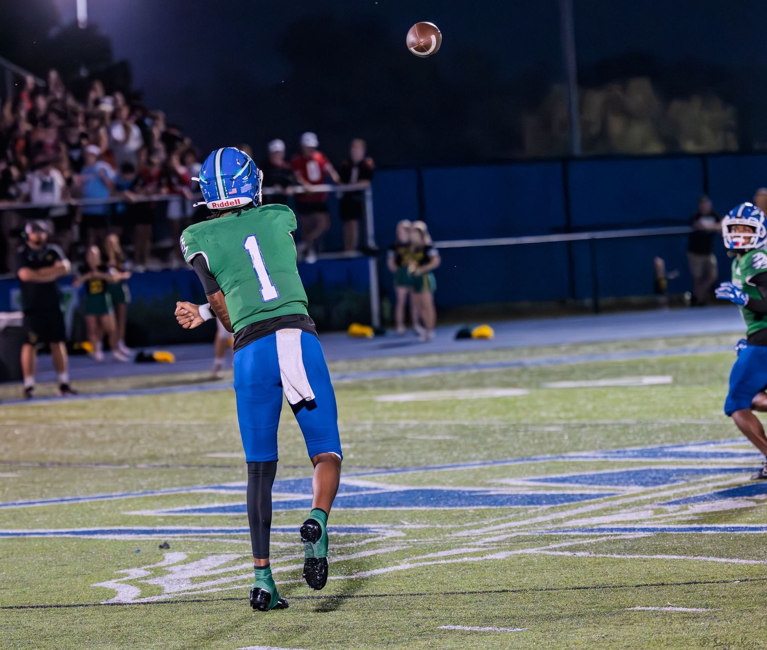 Warriors player number 1 in green uniform throws a pass during a high school football night game.