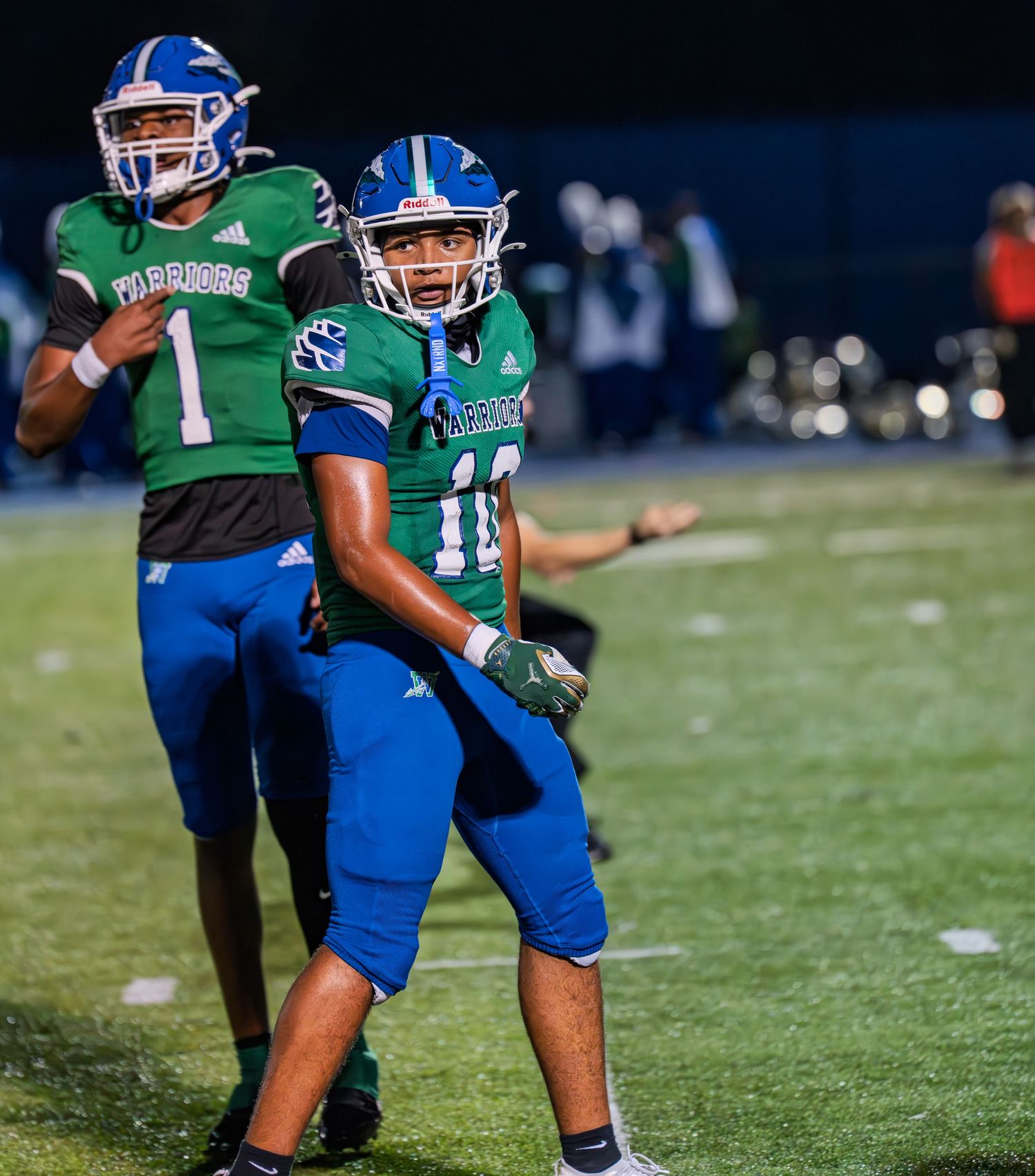 Two Warriors football players in green and blue uniforms celebrate together during a night game.