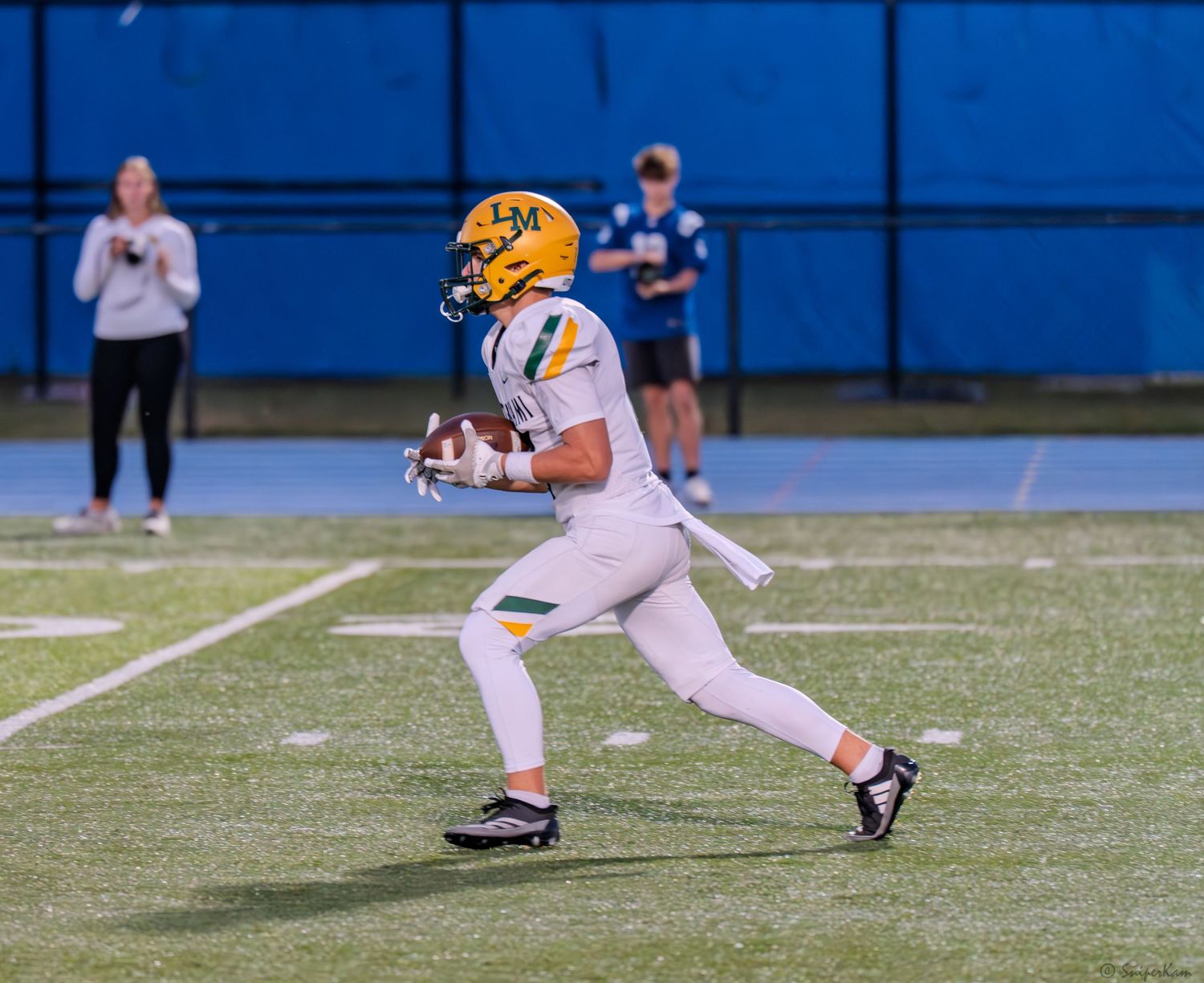 A football player in yellow and white uniform catches a pass during a night game on a blue turf field.