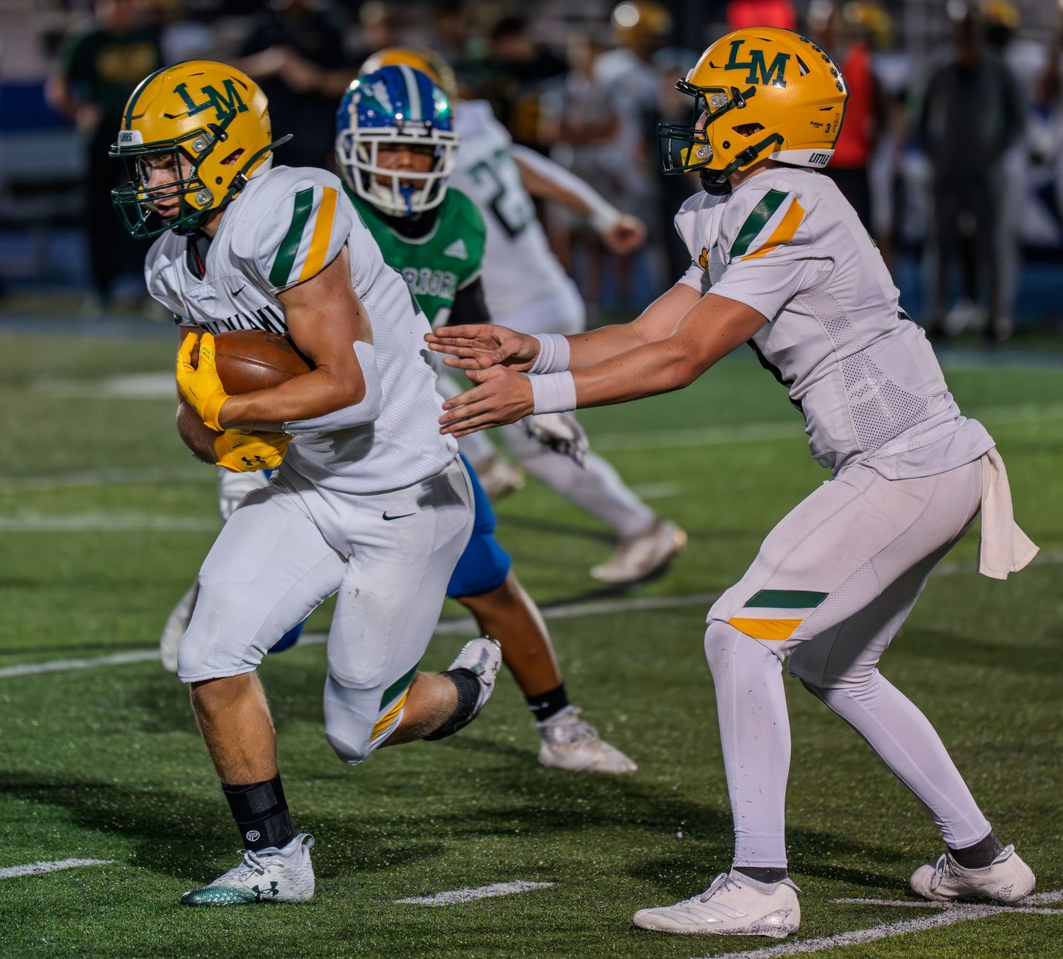 A football player in white and gold uniform receives a handoff from the quarterback during a high school game.