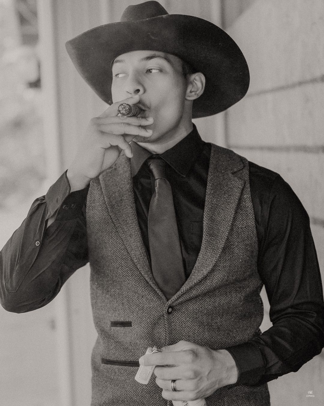 Groom in cowboy hat and tweed vest smoking cigar in black and white wedding portrait.
