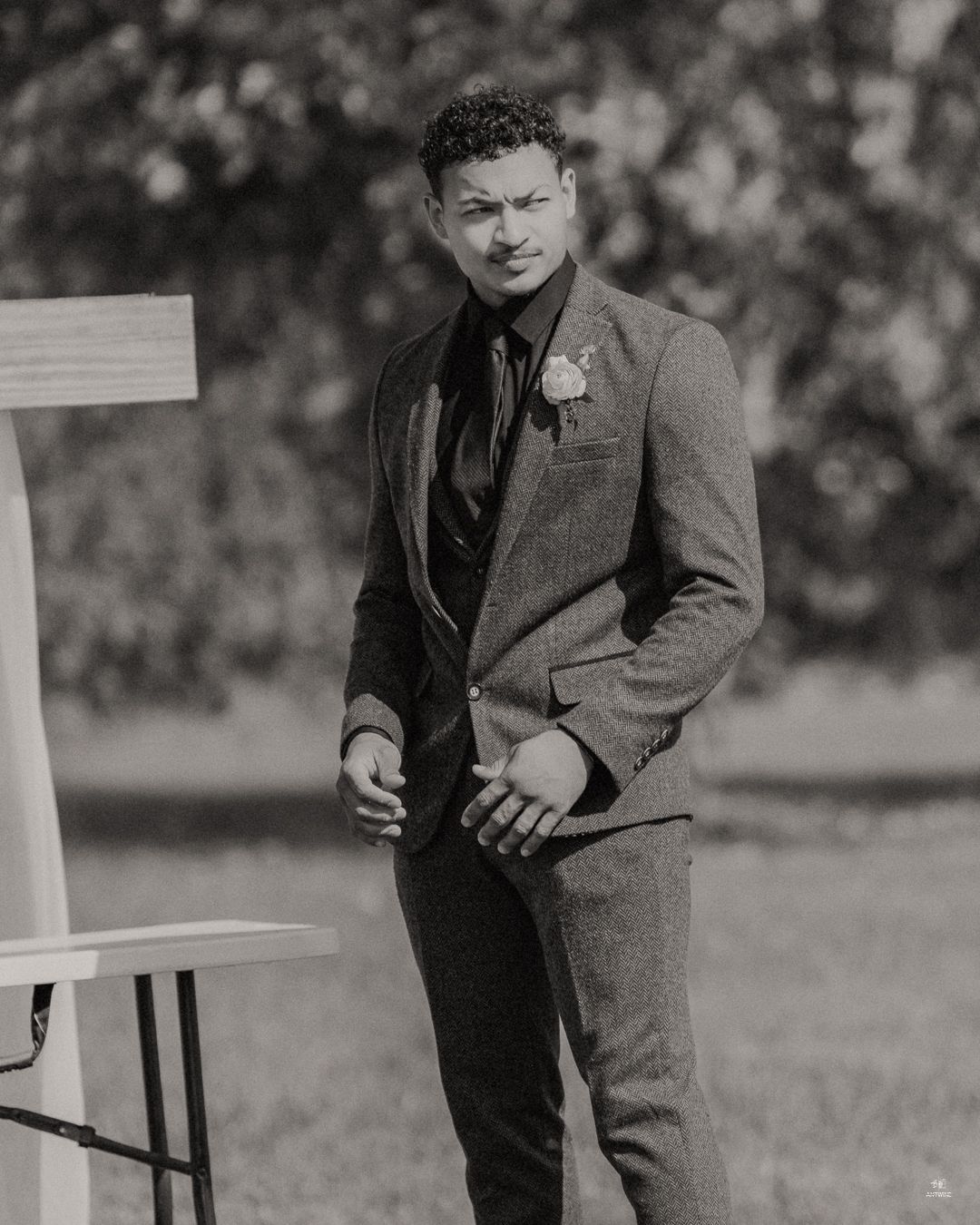 Dapper man in gray herringbone suit with boutonniere poses outdoors at wedding ceremony.