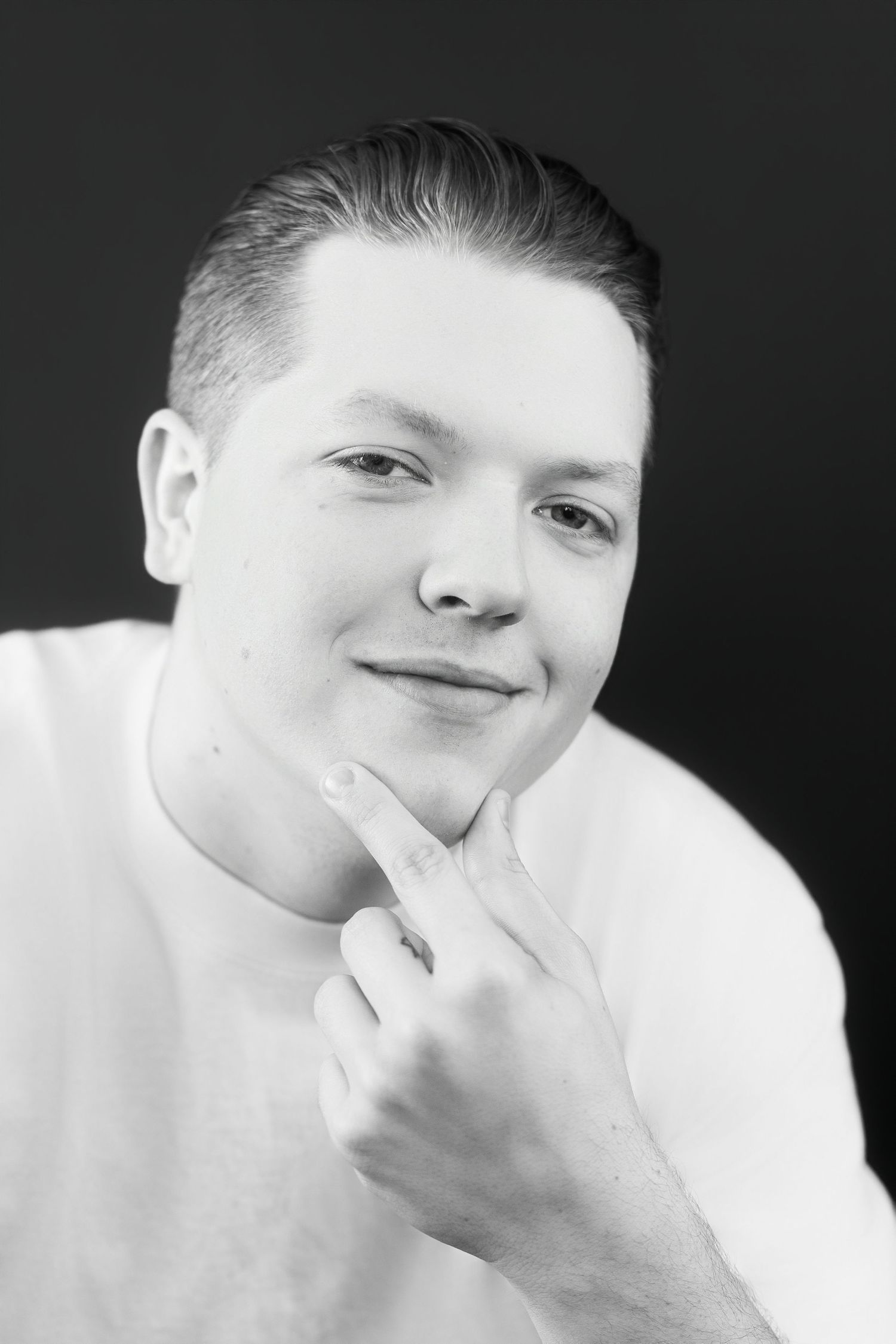 Black and white portrait of smiling young man touching chin with finger against dark background.