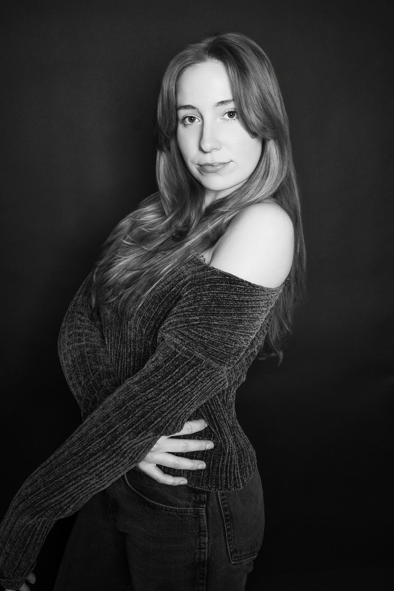 Black and white studio portrait of a young woman in an off-shoulder knit sweater posing gracefully.