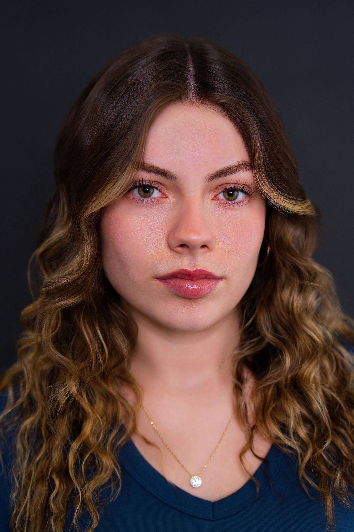 Studio headshot of a young woman with long wavy highlighted hair wearing a blue top and gold necklace.