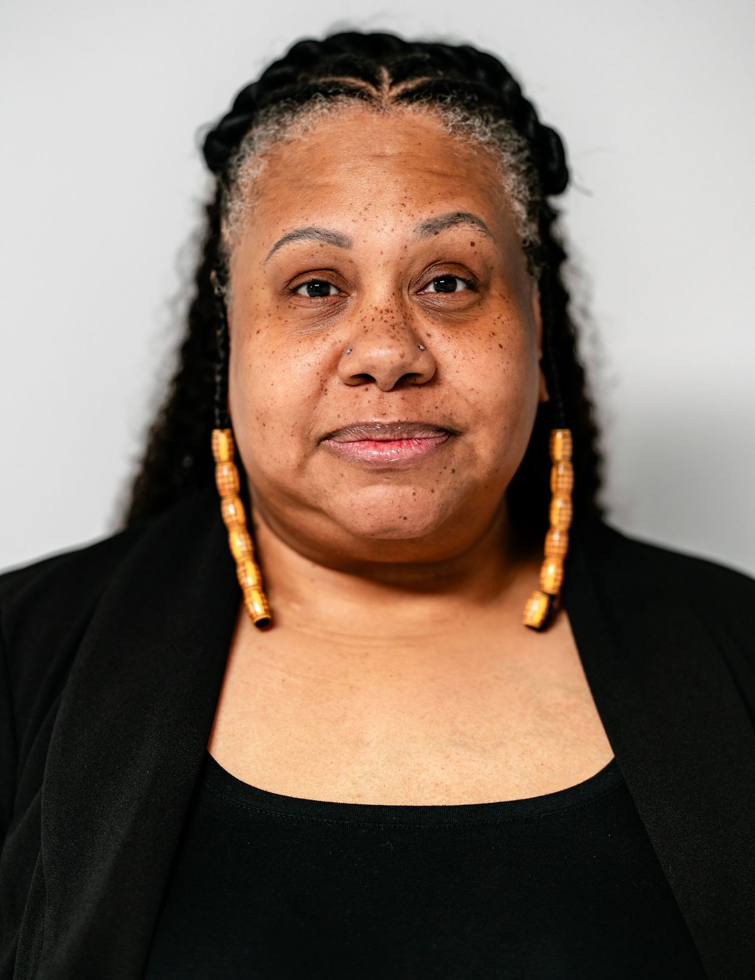 Smiling woman with long braids and gold earrings wearing a black outfit against a light background.