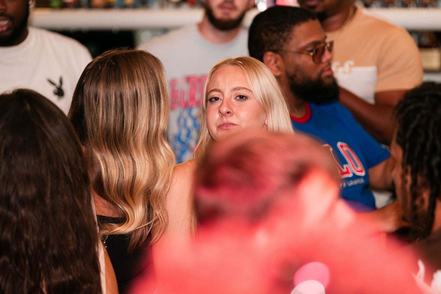 Blonde woman at a crowded indoor social gathering event looking directly at the camera.