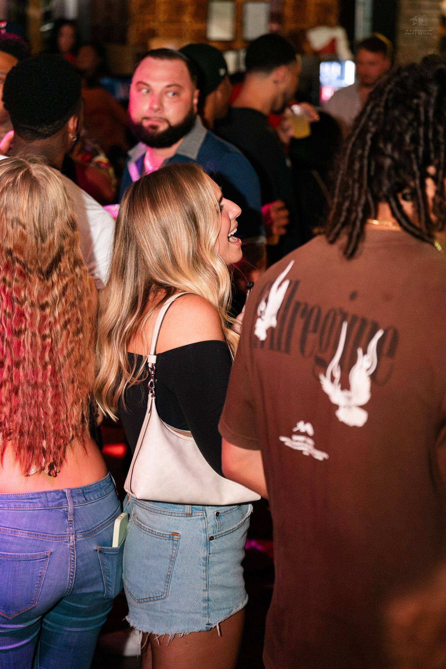 Blonde woman in black off-shoulder top socializing at a packed bar event.