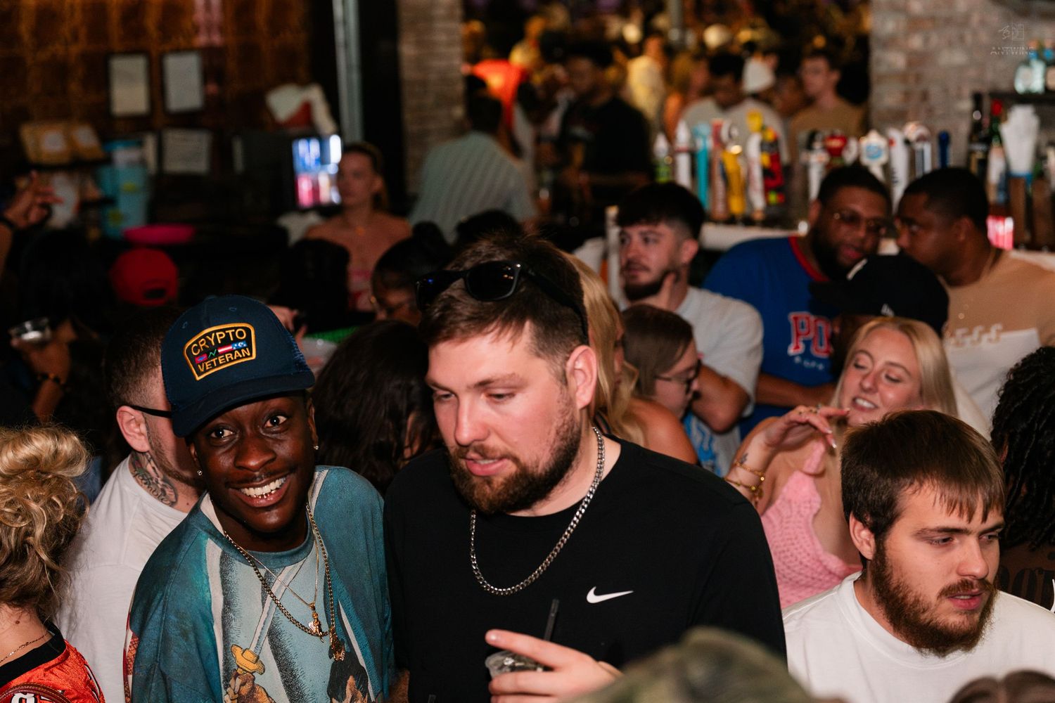 Two men smiling together in a busy crowded bar setting.
