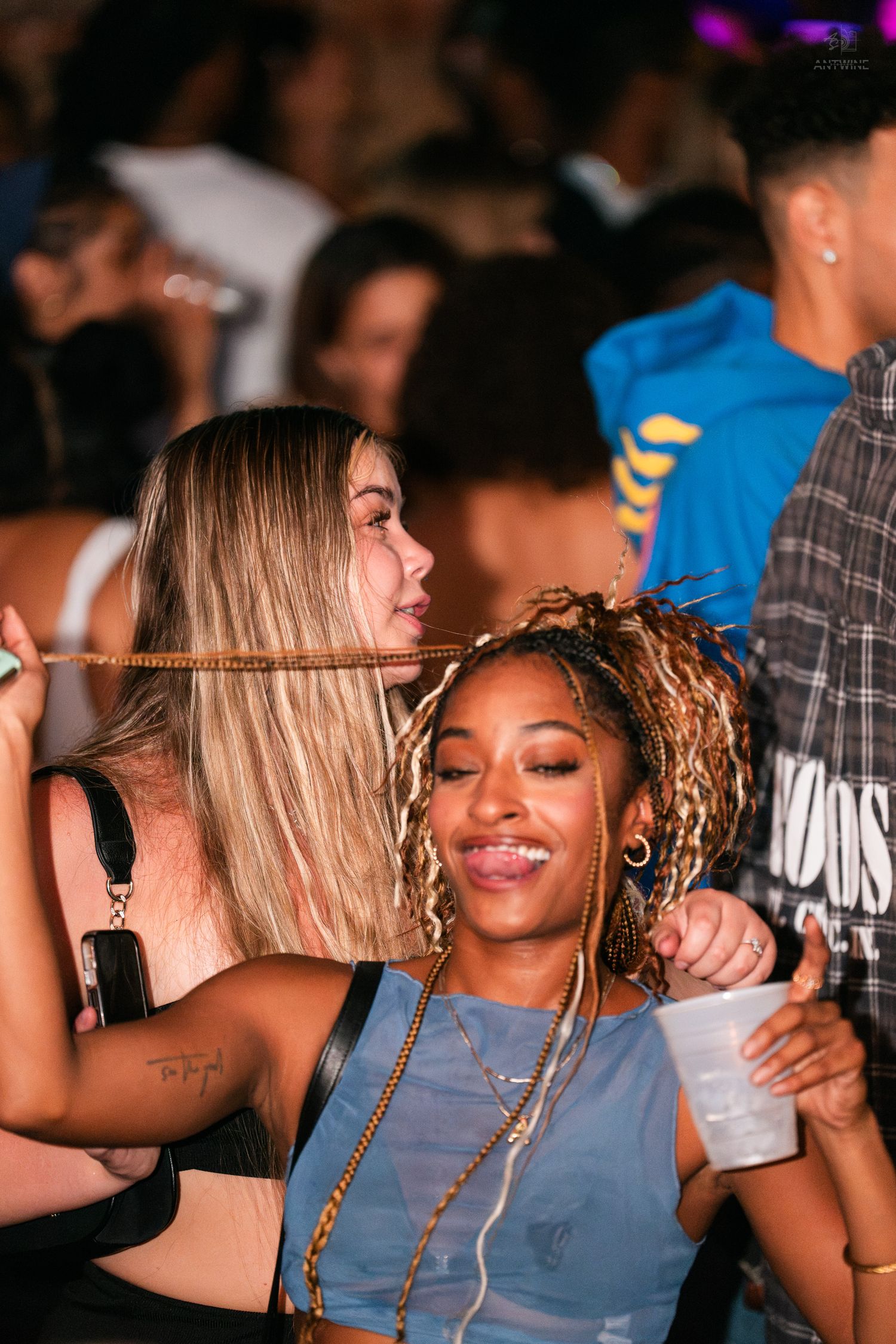 Two women pose together at a nightclub party, one with colorful locs holding a drink.
