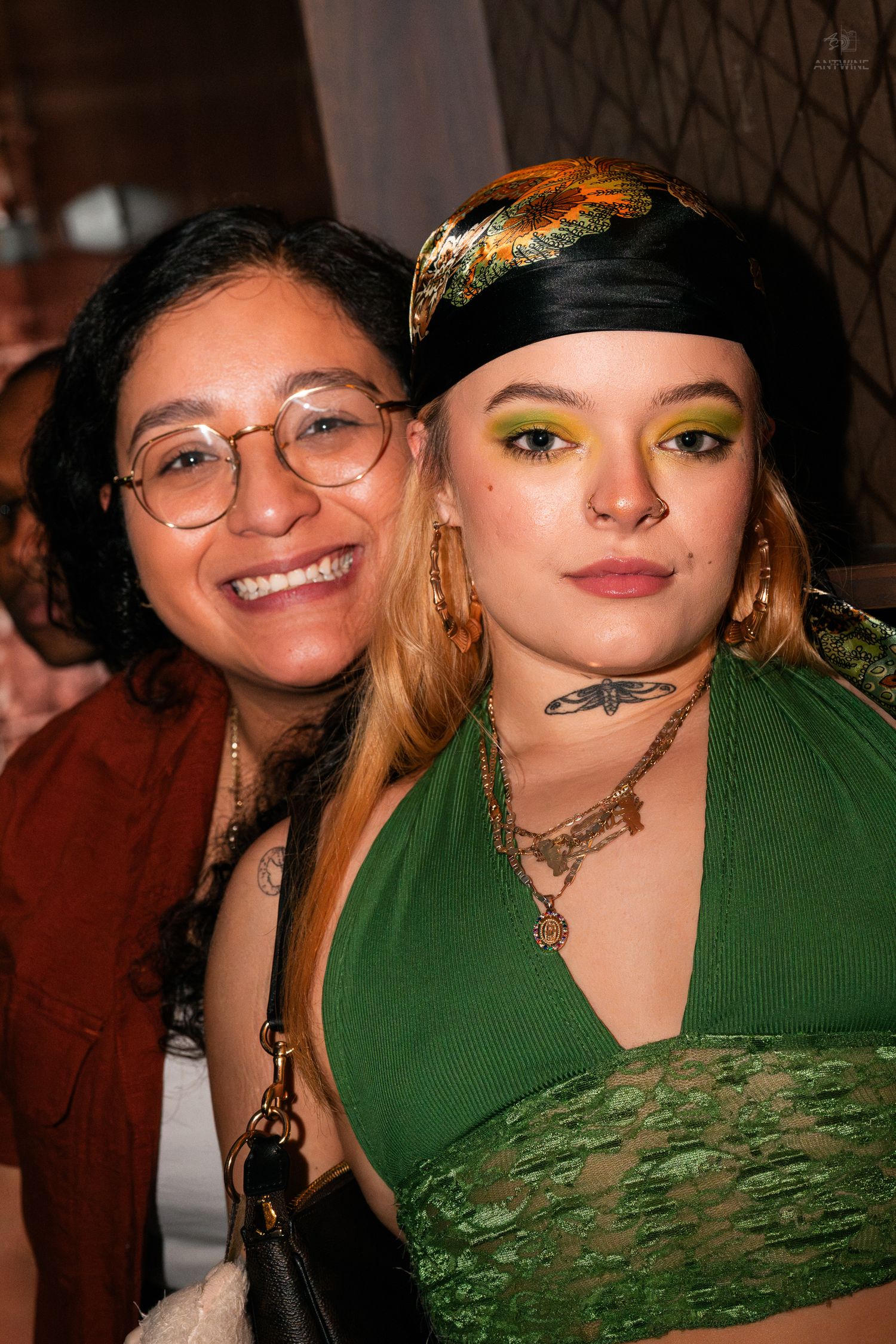 Two women pose together at a party, one wearing bold green eyeshadow and a decorative headband.