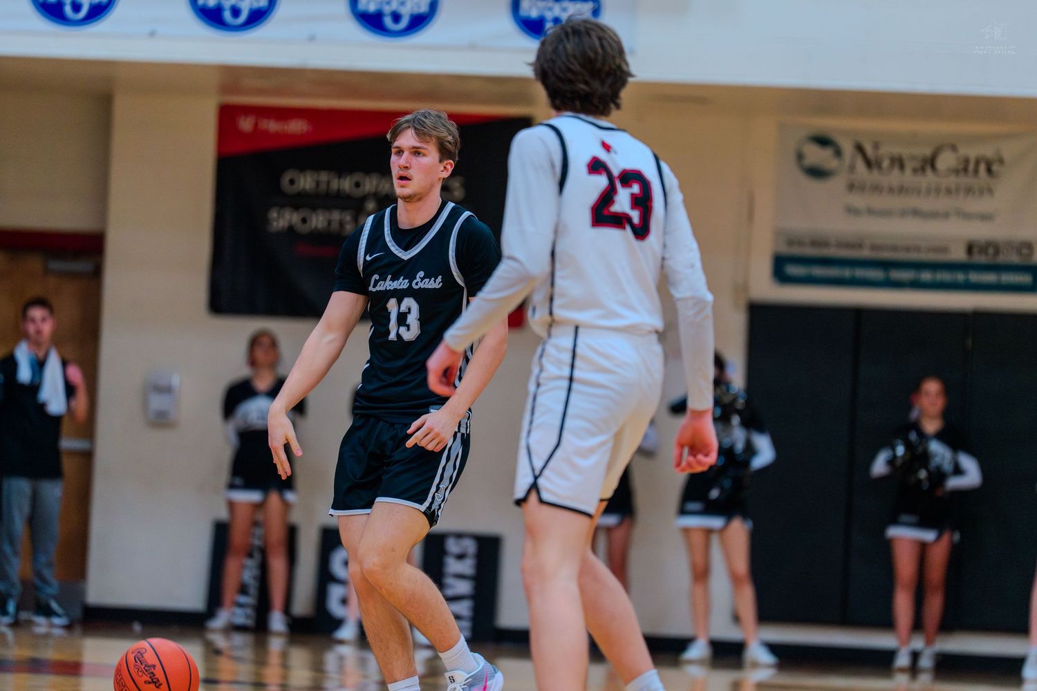 High school basketball player #13 dribbles the ball while defended by opponent #23 during an indoor gym game.