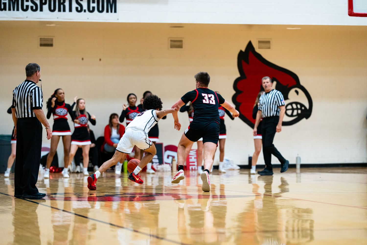 High school basketball players race down court near Cardinals mascot logo during game.