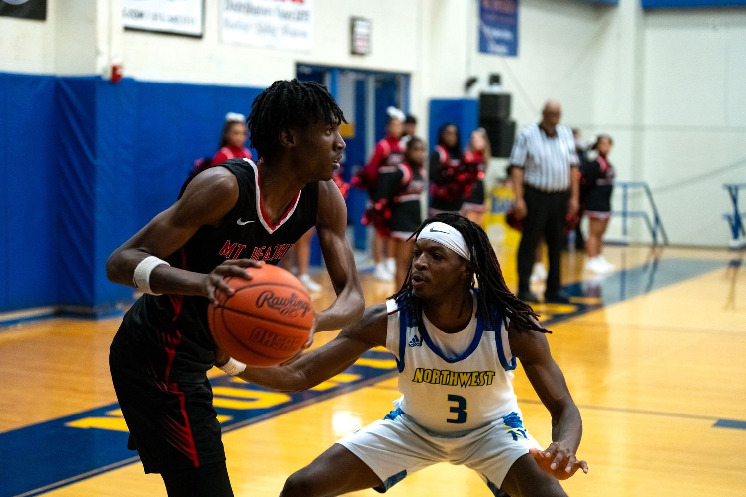 Red jersey player drives baseline while blue jersey defender number 3 contests during basketball game.
