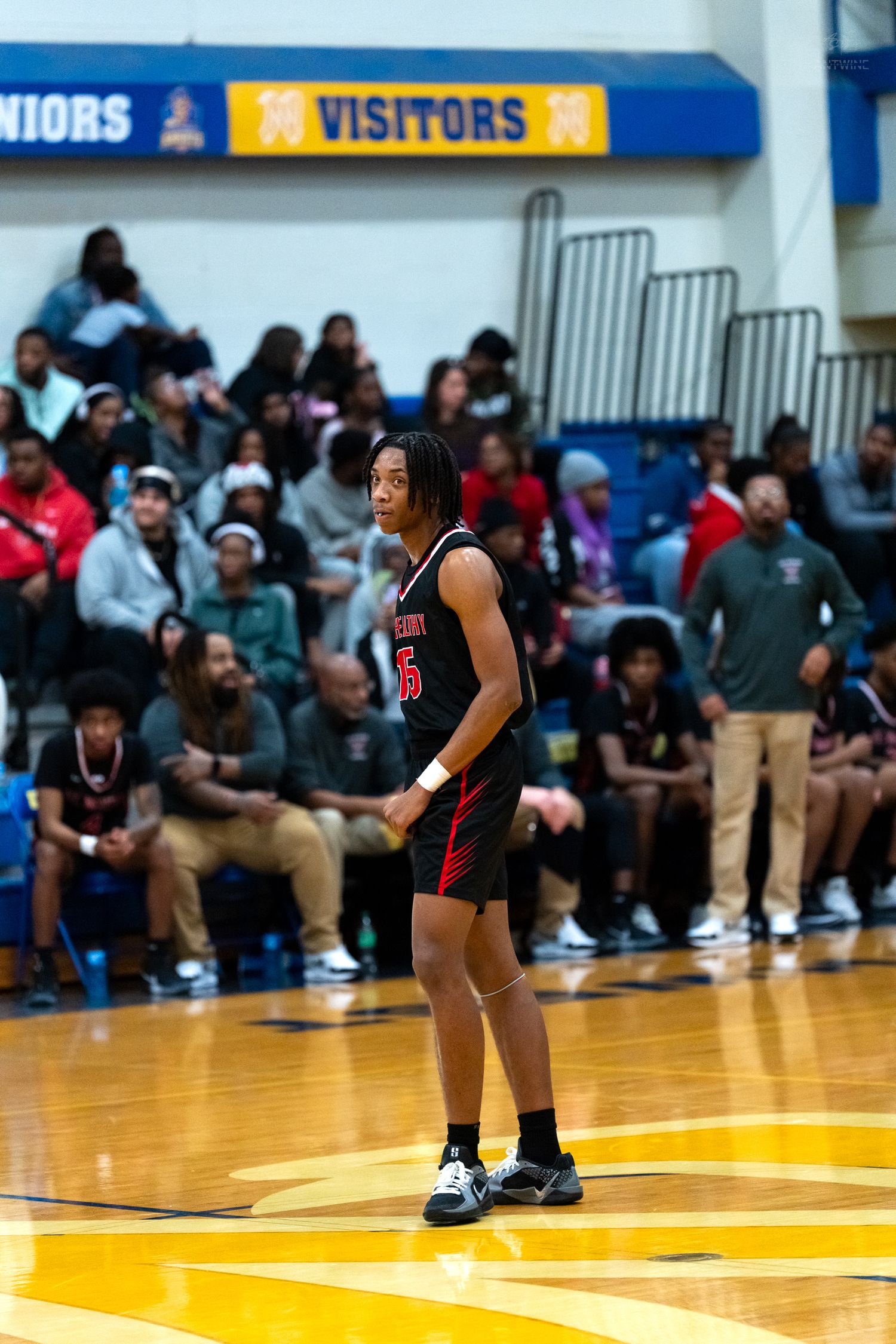 High school basketball player in black and red uniform stands on court with crowd in background.