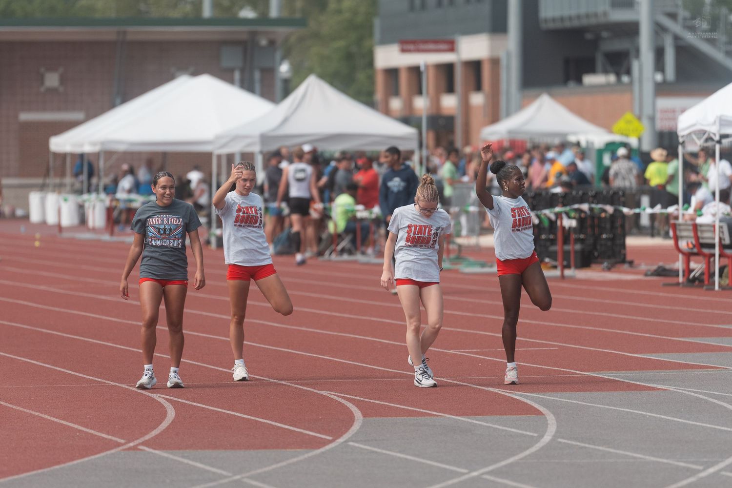 Four female track athletes in matching red uniforms jog along a red track during a field day competition.