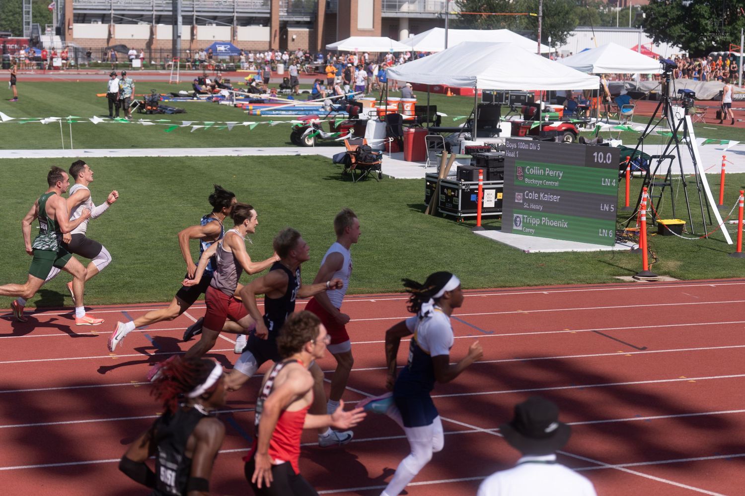 A large group of runners compete in a distance race on a red track during an outdoor track and field meet.