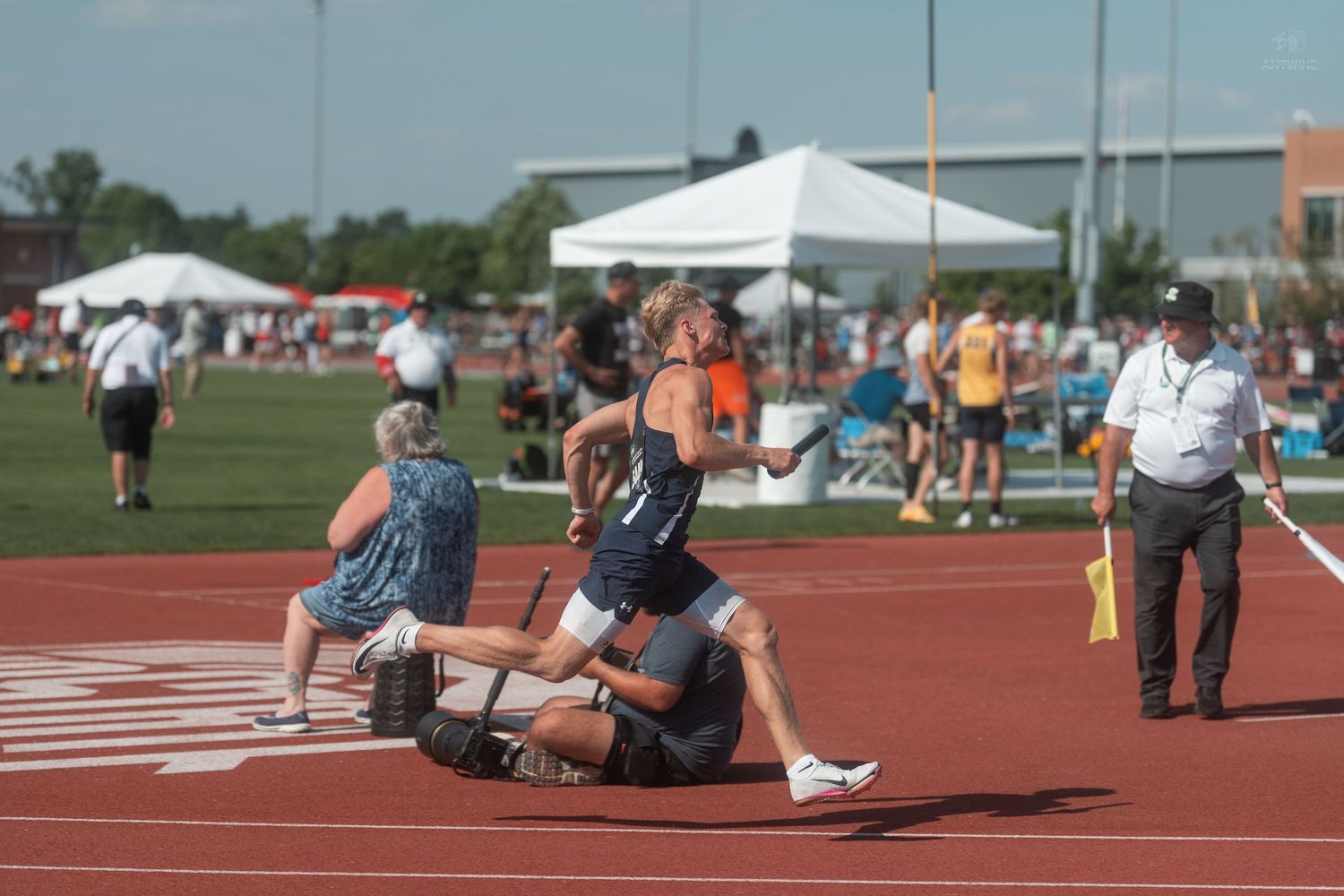 Athletes compete in a relay race on a red track during an outdoor track and field meet with spectators nearby.