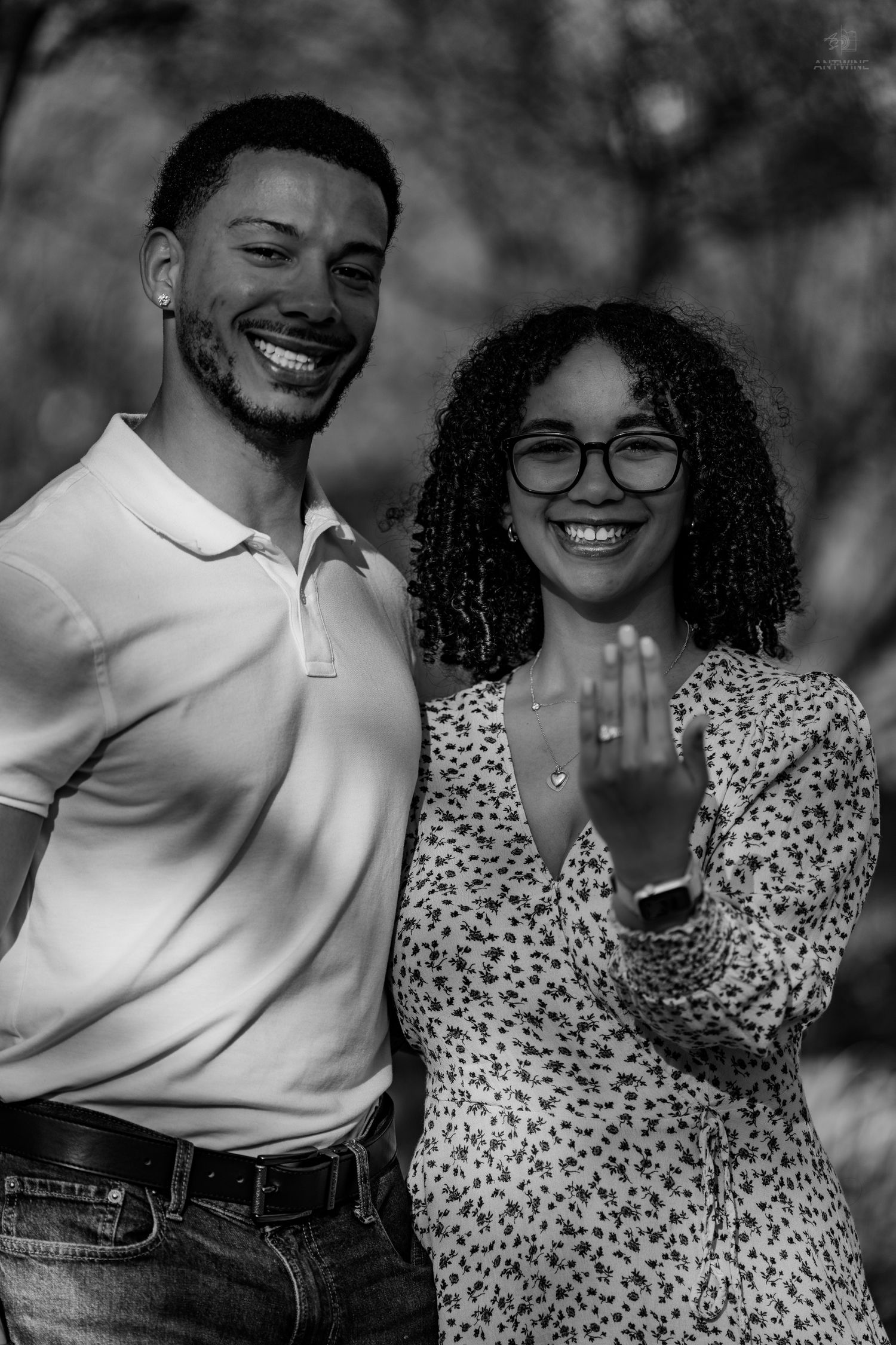 Black and white photo of smiling engaged couple, woman proudly displaying her new engagement ring outdoors.